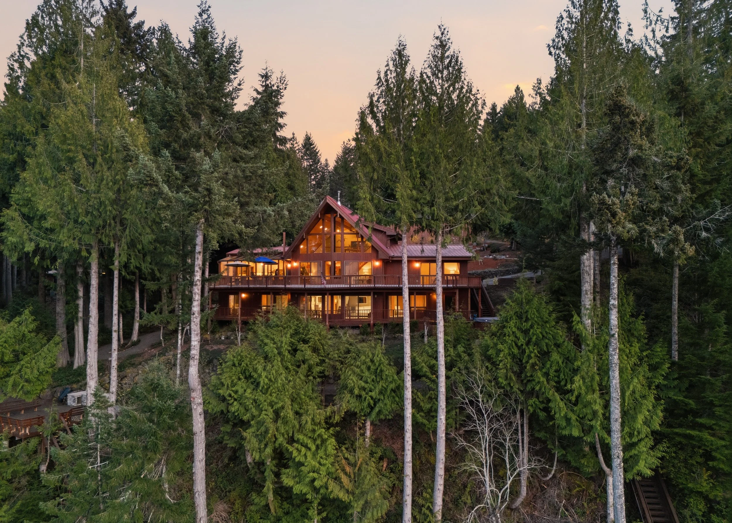 A large wooden house with multiple floors and large windows, illuminated from inside, situated on a forested hillside surrounded by tall pine trees at dusk.