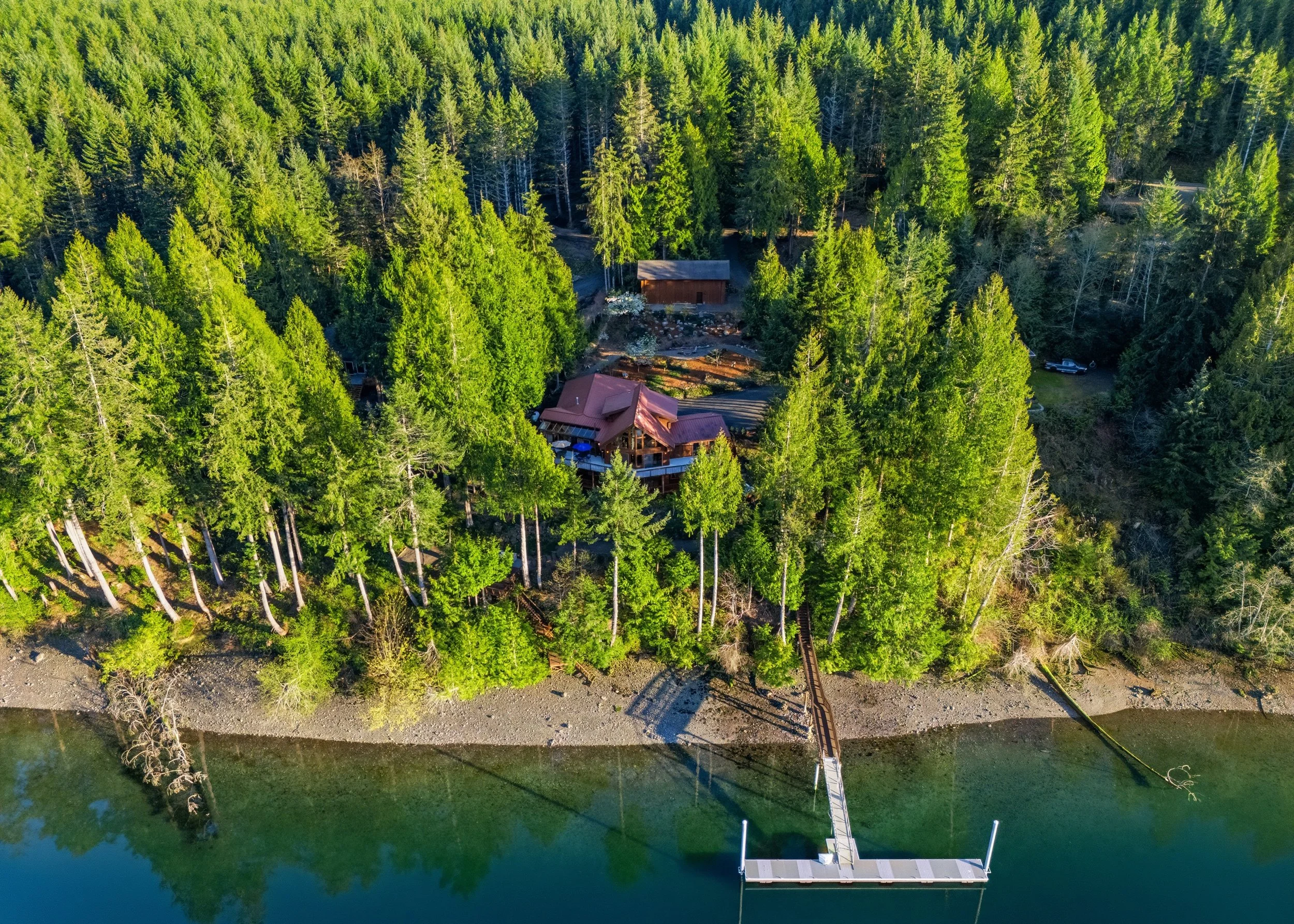 Aerial view of a house surrounded by tall evergreen trees, located on a shoreline with a dock extending into the water.