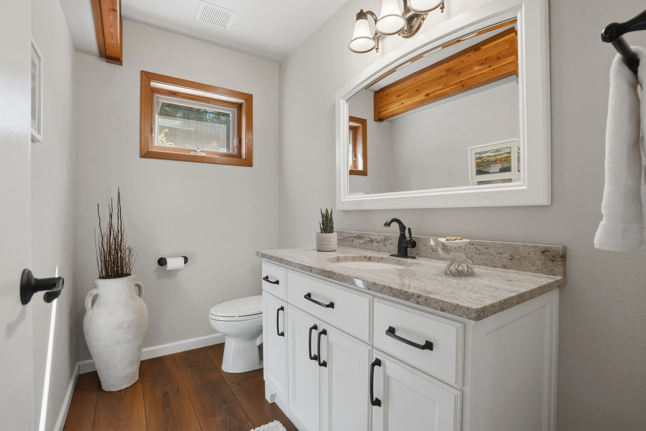A bathroom with a white vanity and granite countertop, a black faucet, a large mirror, two small windows with wood trim, a potted plant, and a vase with decorative twigs.