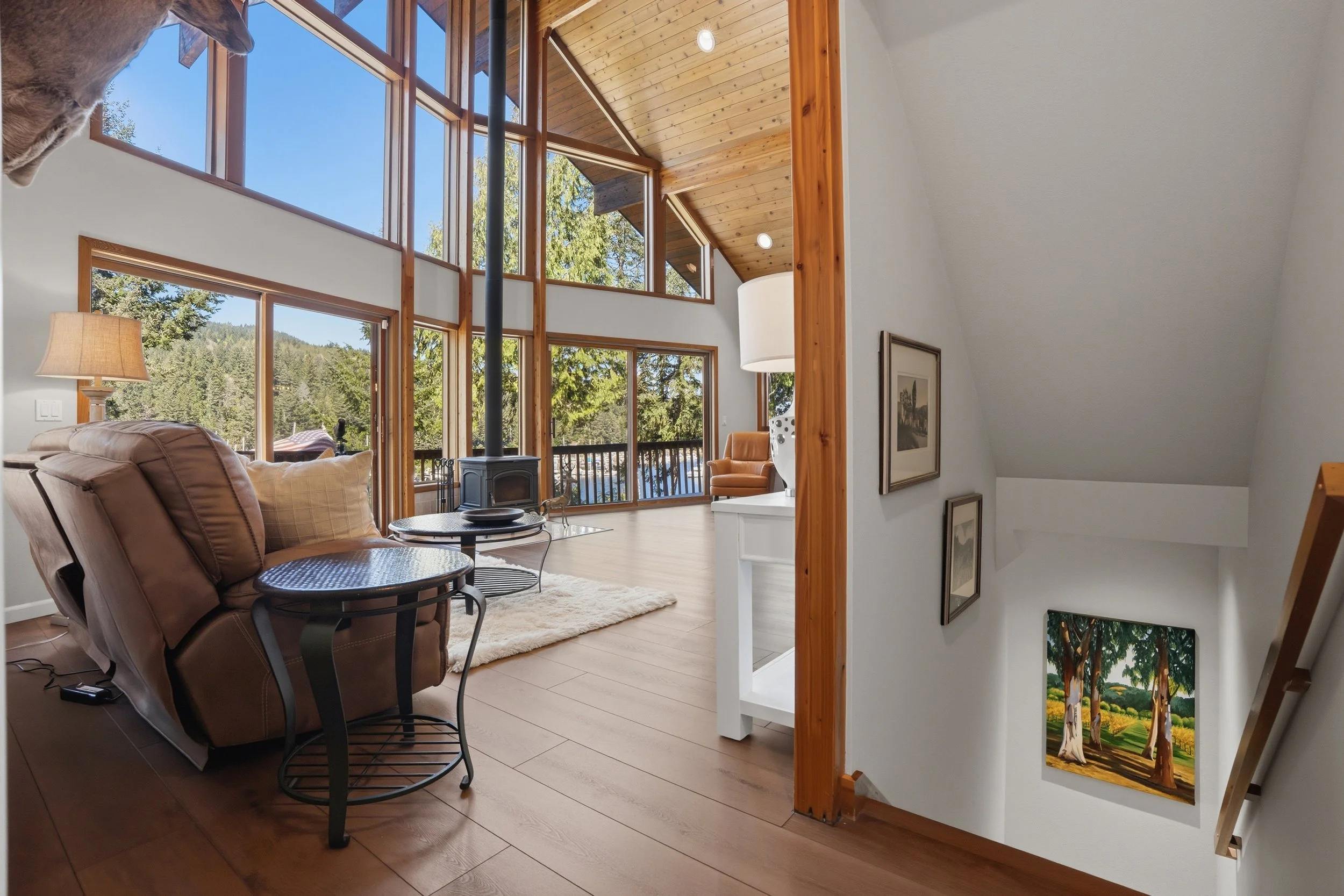 Living room with large windows, wooden ceiling, and a mountain view. Features a brown recliner, a small black metal side table, a white fireplace, and an area rug. The staircase leads down to a lower level with framed artwork on the wall.