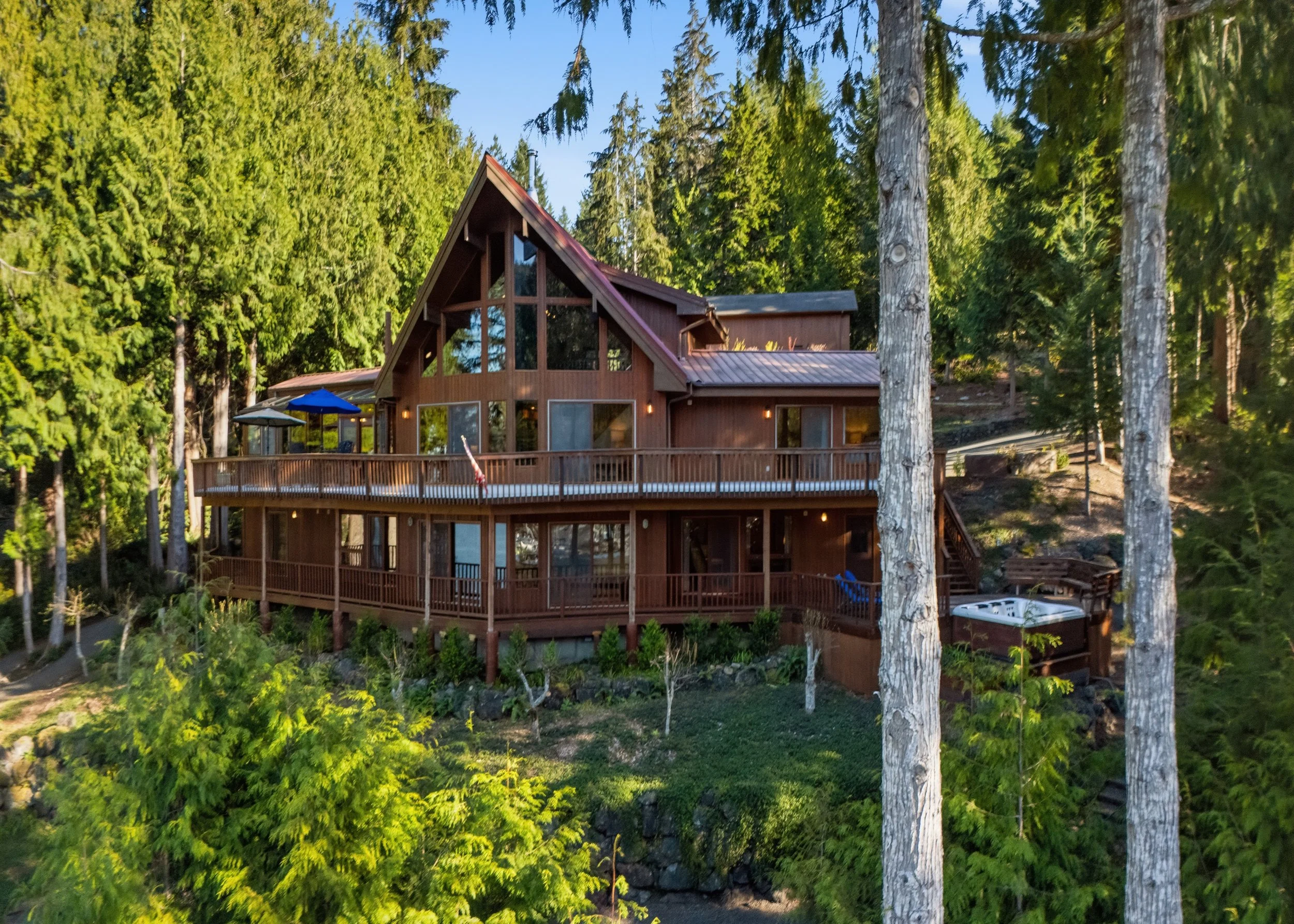 A large wooden house surrounded by lush green trees, with a spacious elevated deck and outdoor hot tub.