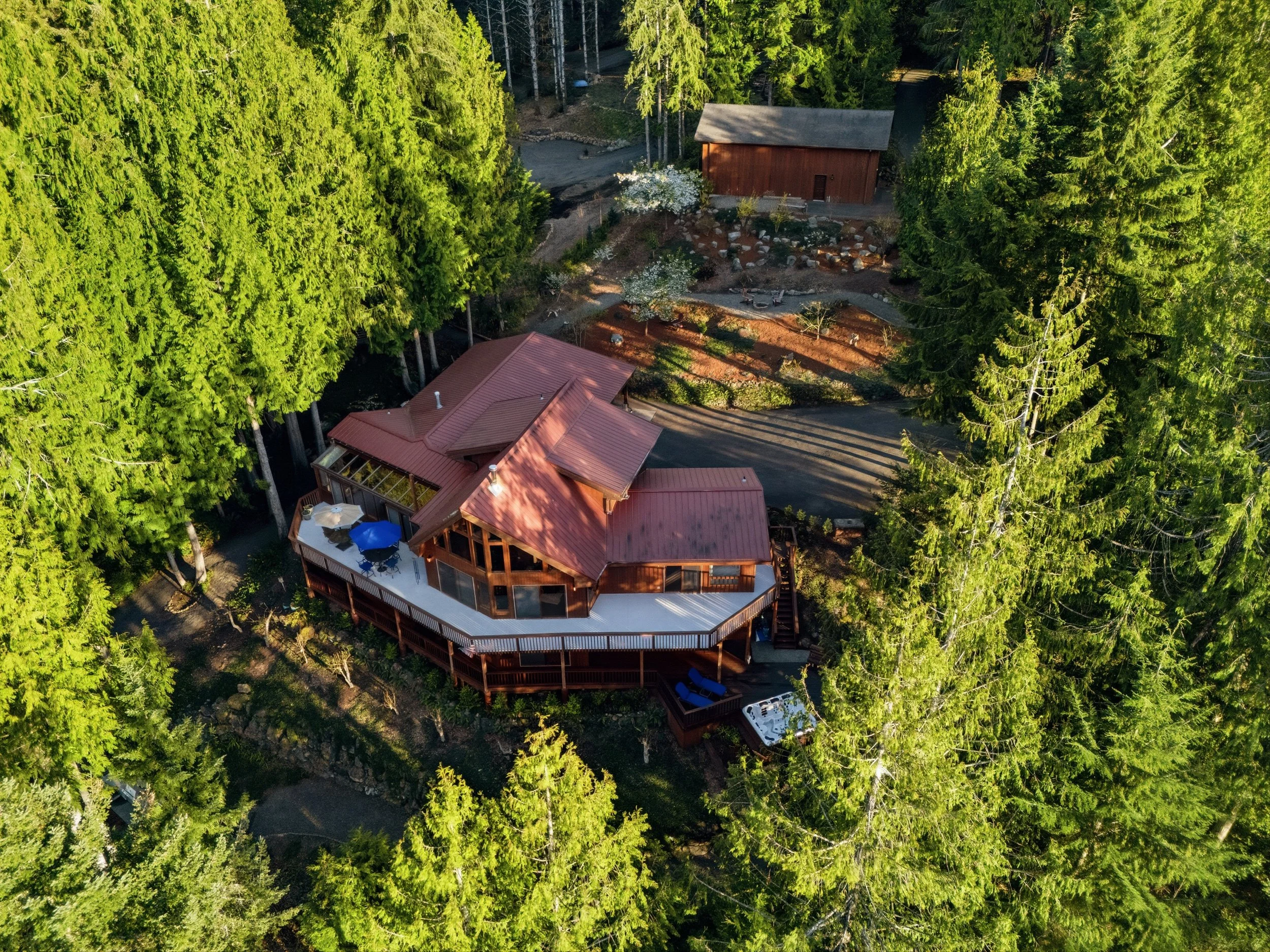 A house with a large deck surrounded by tall green trees, with outdoor furniture and a hot tub visible.