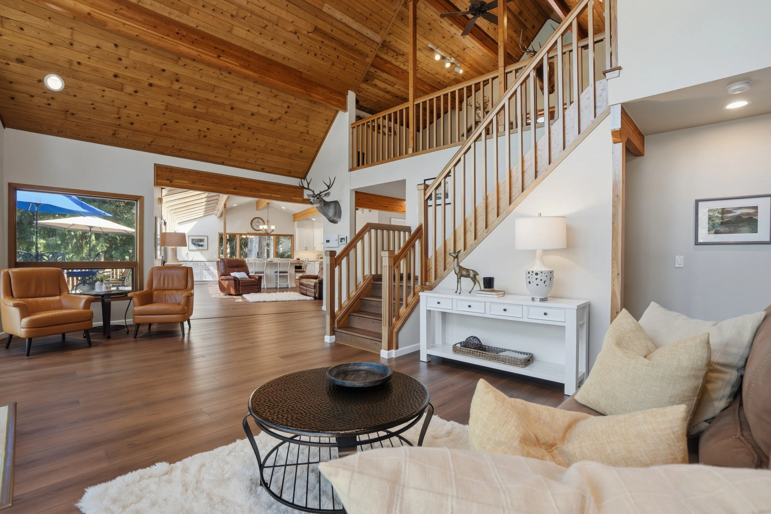 Living room with wooden ceiling and staircase, tan leather chairs, white decorated side table with lamp, beige sofa with pillows, large window showing outdoor patio with umbrella, and a mounted deer head on the wall.