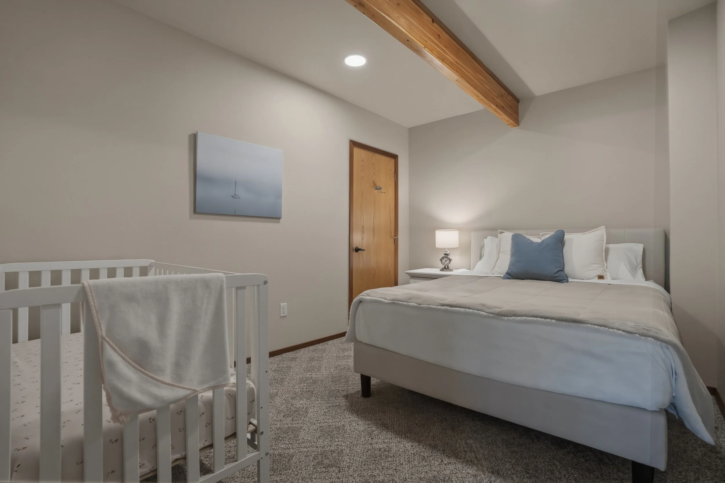 A bedroom with a large bed, nightstand with lamp, and a crib in the corner, with a wooden ceiling beam and framed picture on the wall.