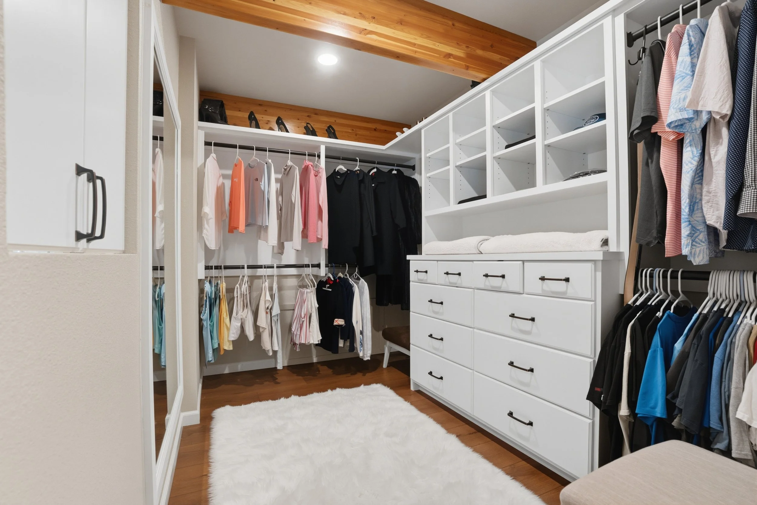 Organized walk-in closet with hanging clothes, shelves, and a white dresser with towels, featuring a wooden ceiling and hardwood floor.
