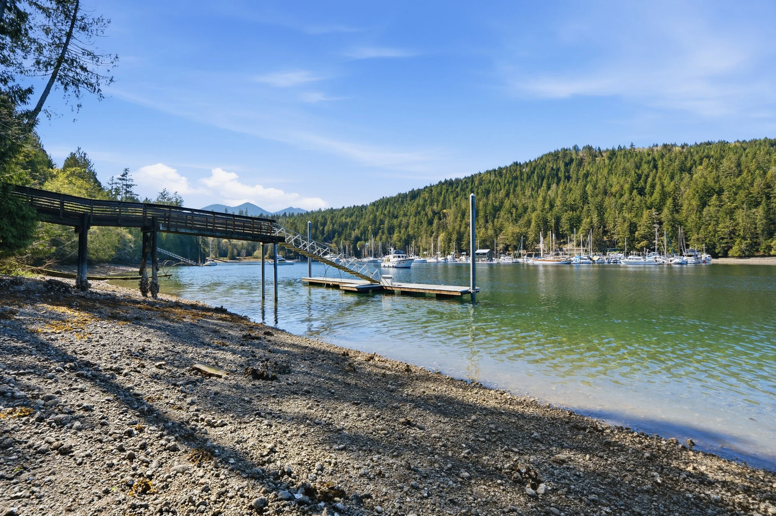 A gravel shore next to a body of water with a small dock and boat ramp, surrounded by a forested hillside, under a partly cloudy sky.