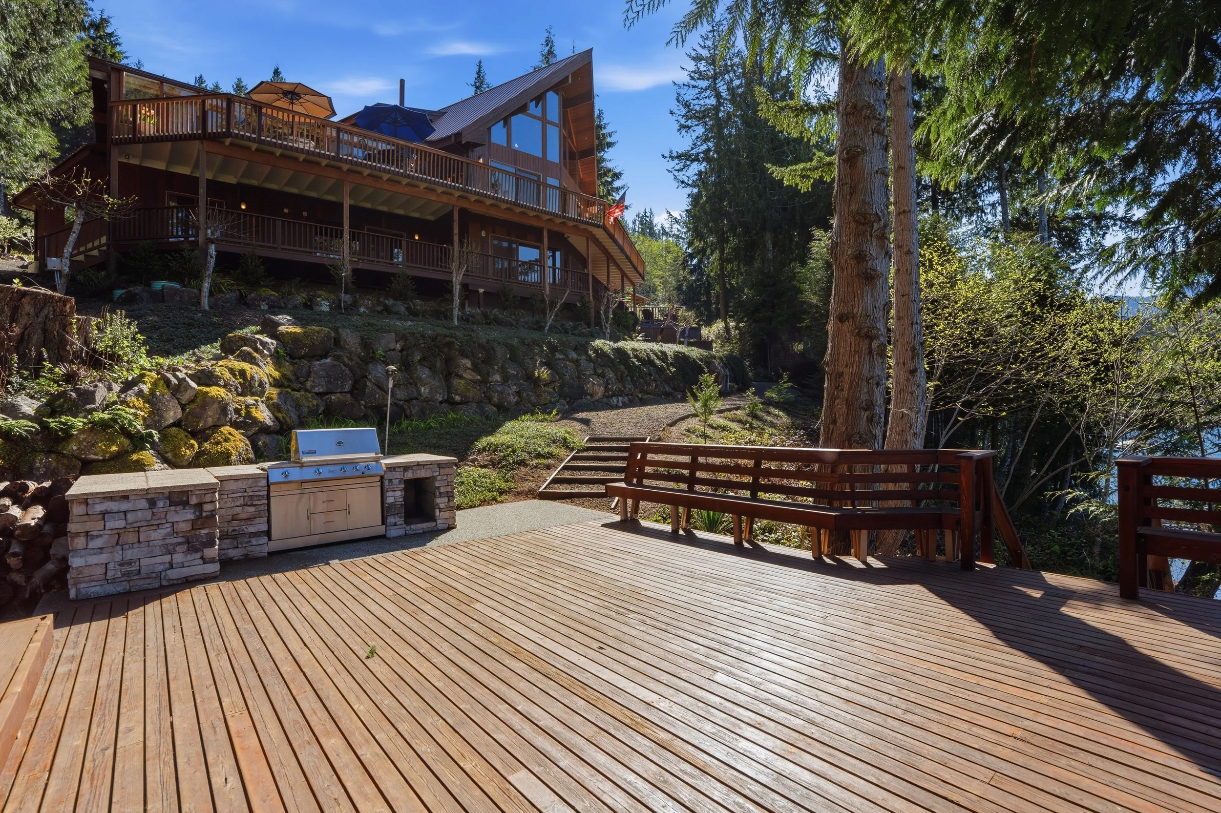 Wooden deck with built-in grill in a backyard surrounded by trees and a hillside with a large house in the background