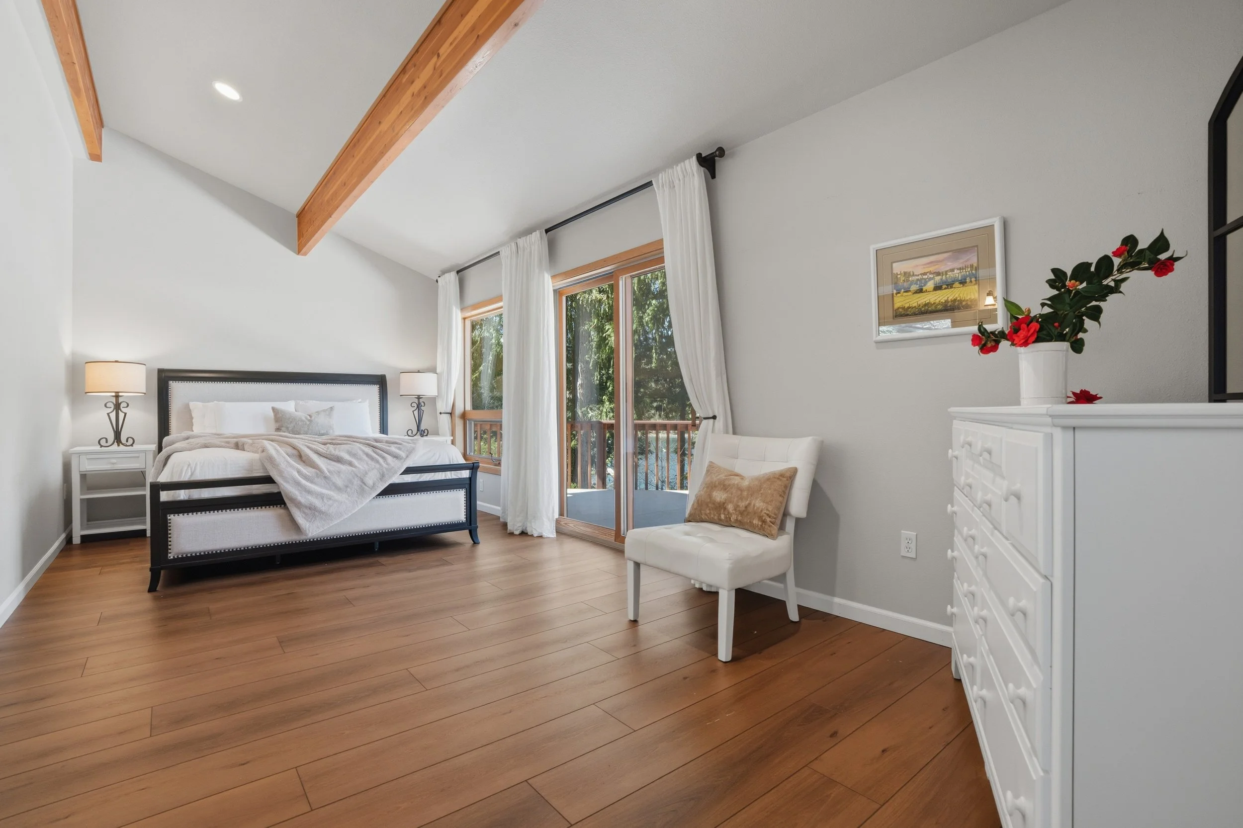 Bright bedroom with wooden floors, a large bed with white bedding, white nightstands with lamps, a white chair with a tan pillow, glass sliding door to a balcony, and a white dresser with a red flower vase.