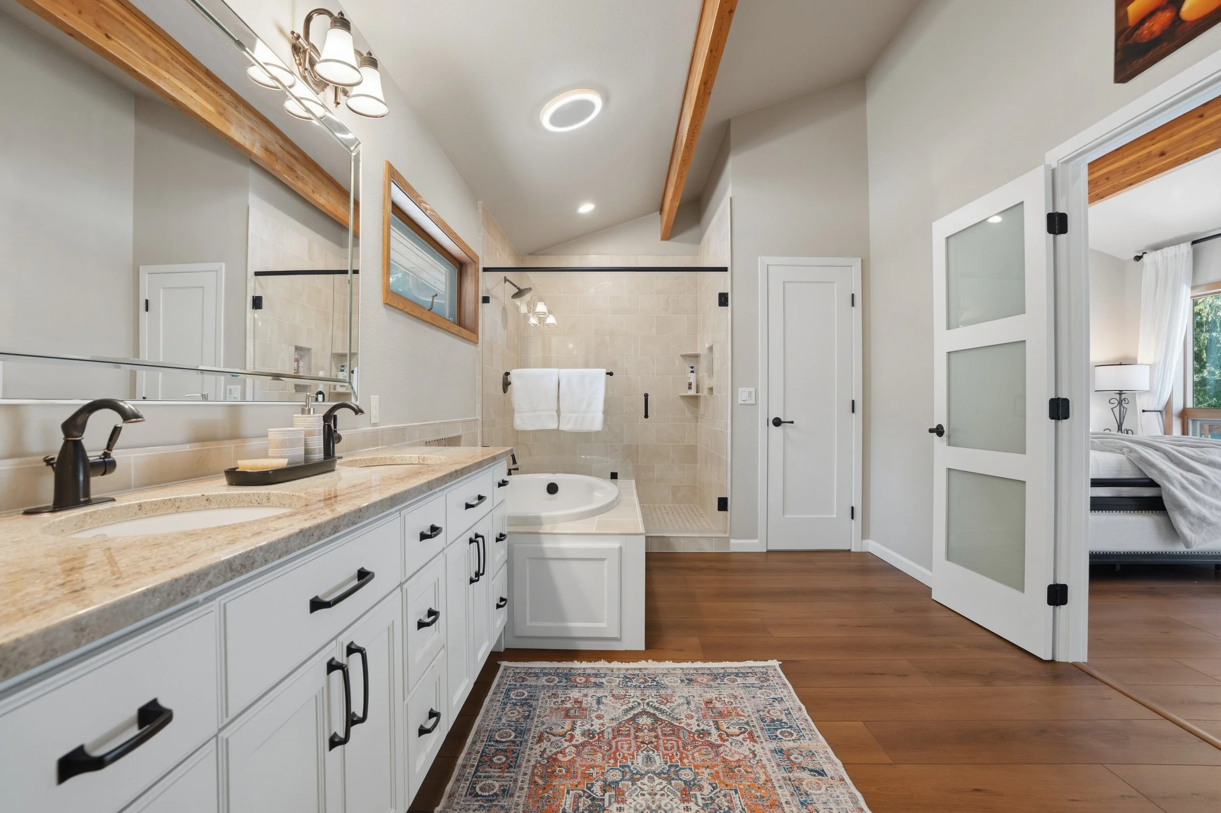 Modern bathroom with white cabinetry, double sink with black fixtures, beige countertop, large mirrored wall, and walk-in shower with glass door. Bedroom visible through sliding door with white curtains.