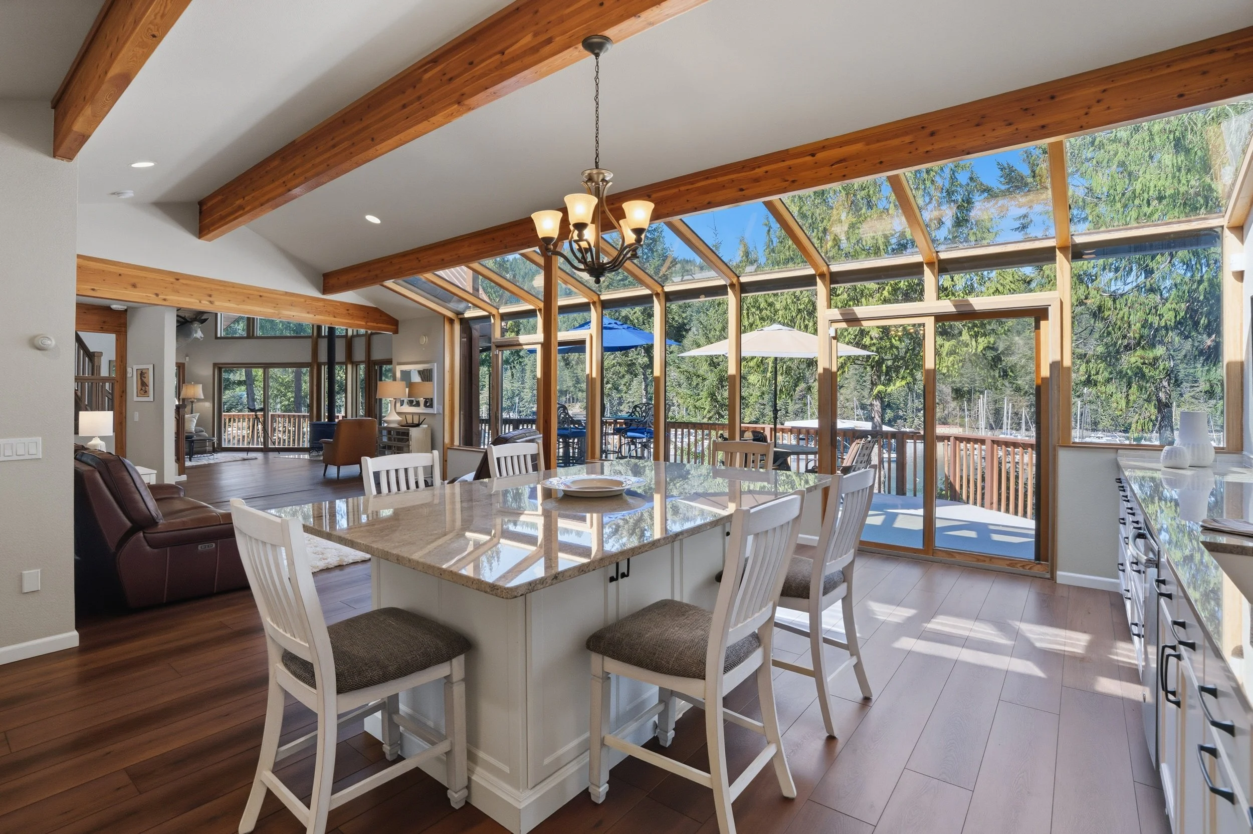 Open-concept living and dining area with large glass windows and sliding door, wood beams, and a deck outside with umbrellas and outdoor furniture.