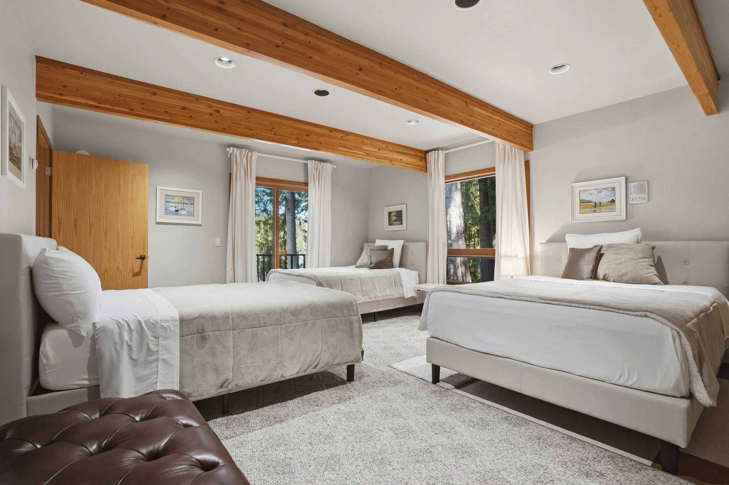 A spacious bedroom with three white beds, two larger beds on the right and one smaller on the left. Light-colored walls, framed artwork above each bed, large windows with white curtains, and wooden ceiling beams.