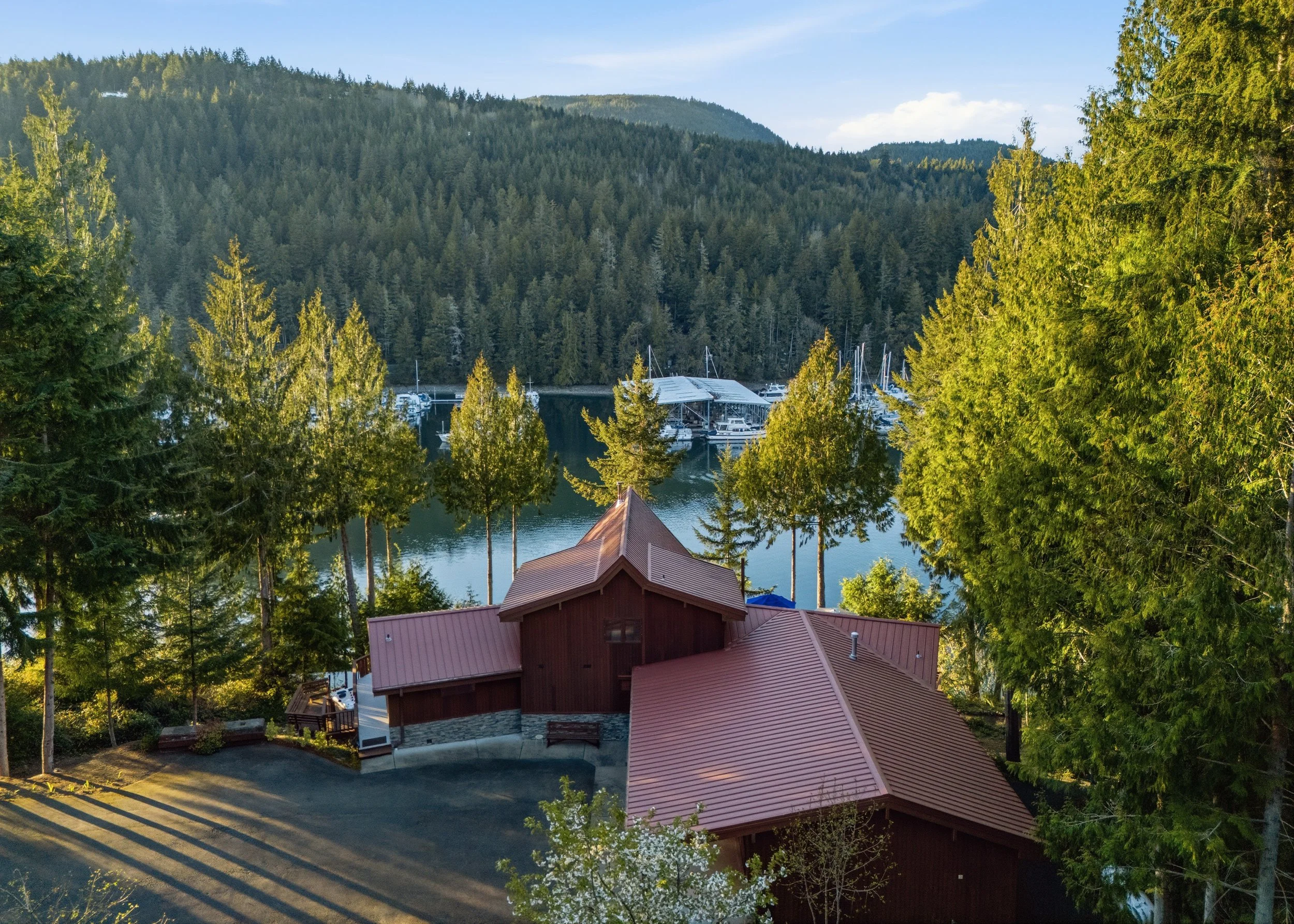 A house with a red roof situated among green trees on a hillside overlooking a lake with boats and a forested mountain in the background.