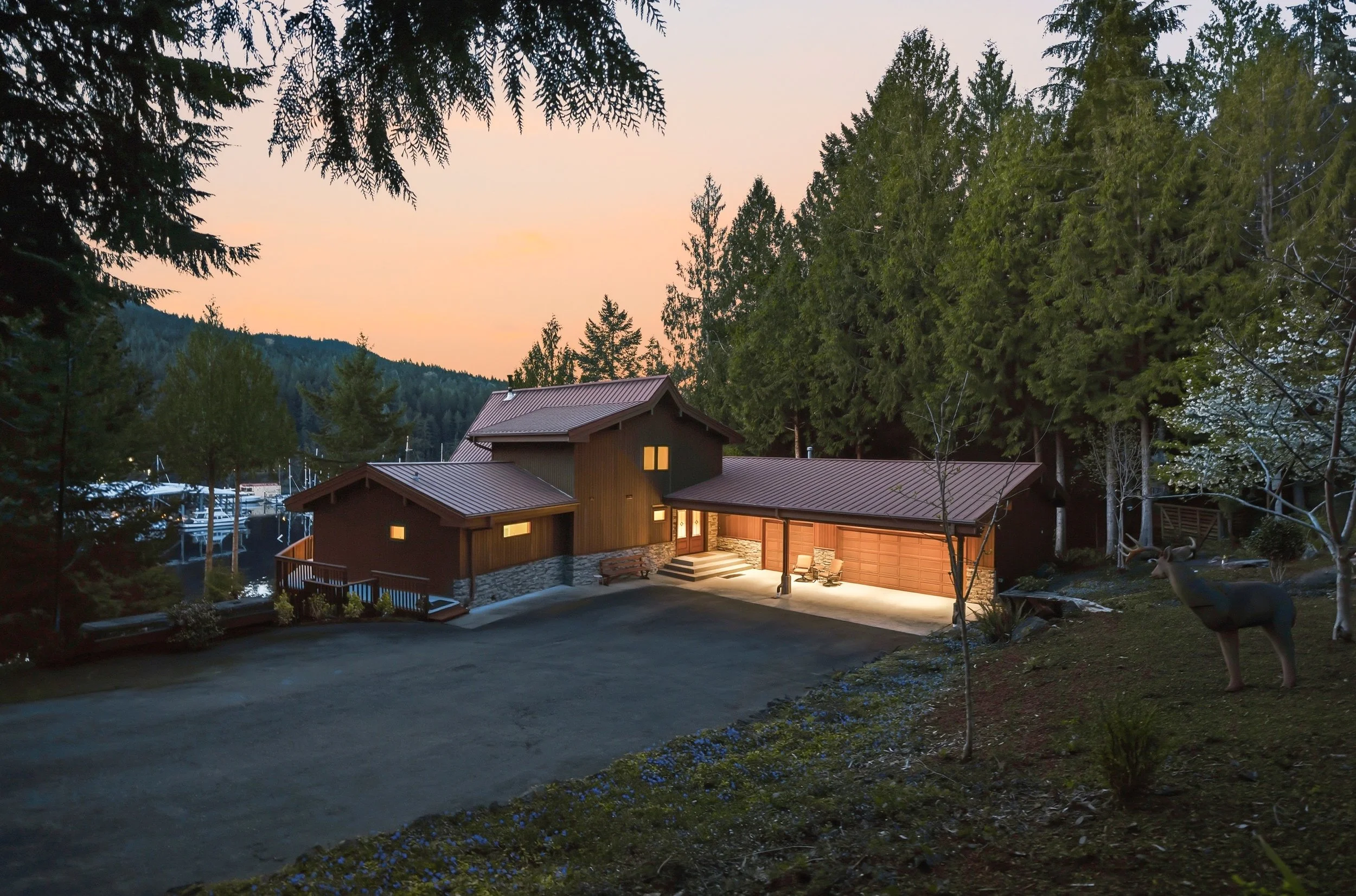 A modern house made of wood and stone, illuminated at dusk, nestled among tall trees and with a driveway in front.