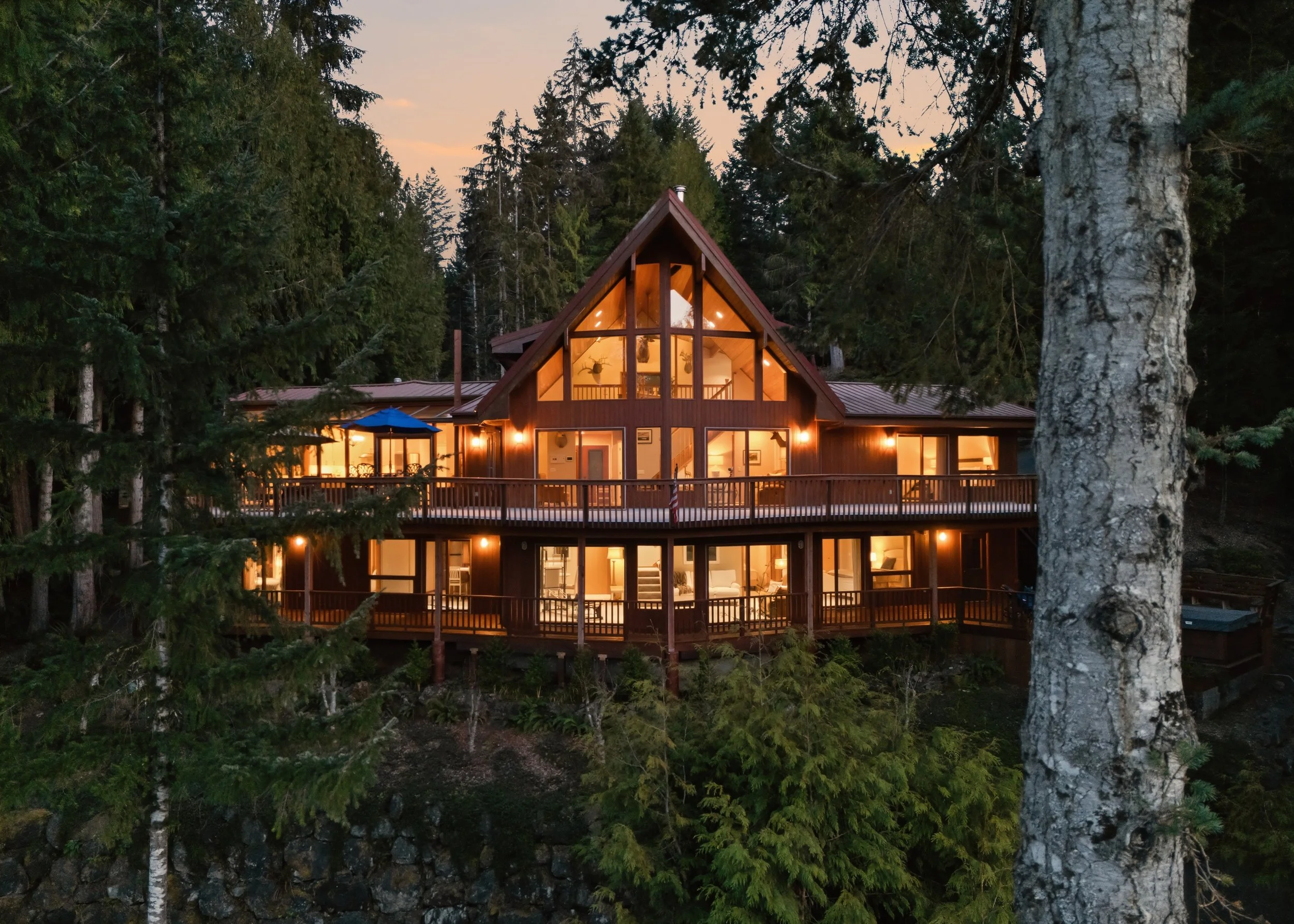 A large A-frame house with multiple levels, illuminated from within, set among tall pine trees at dusk.