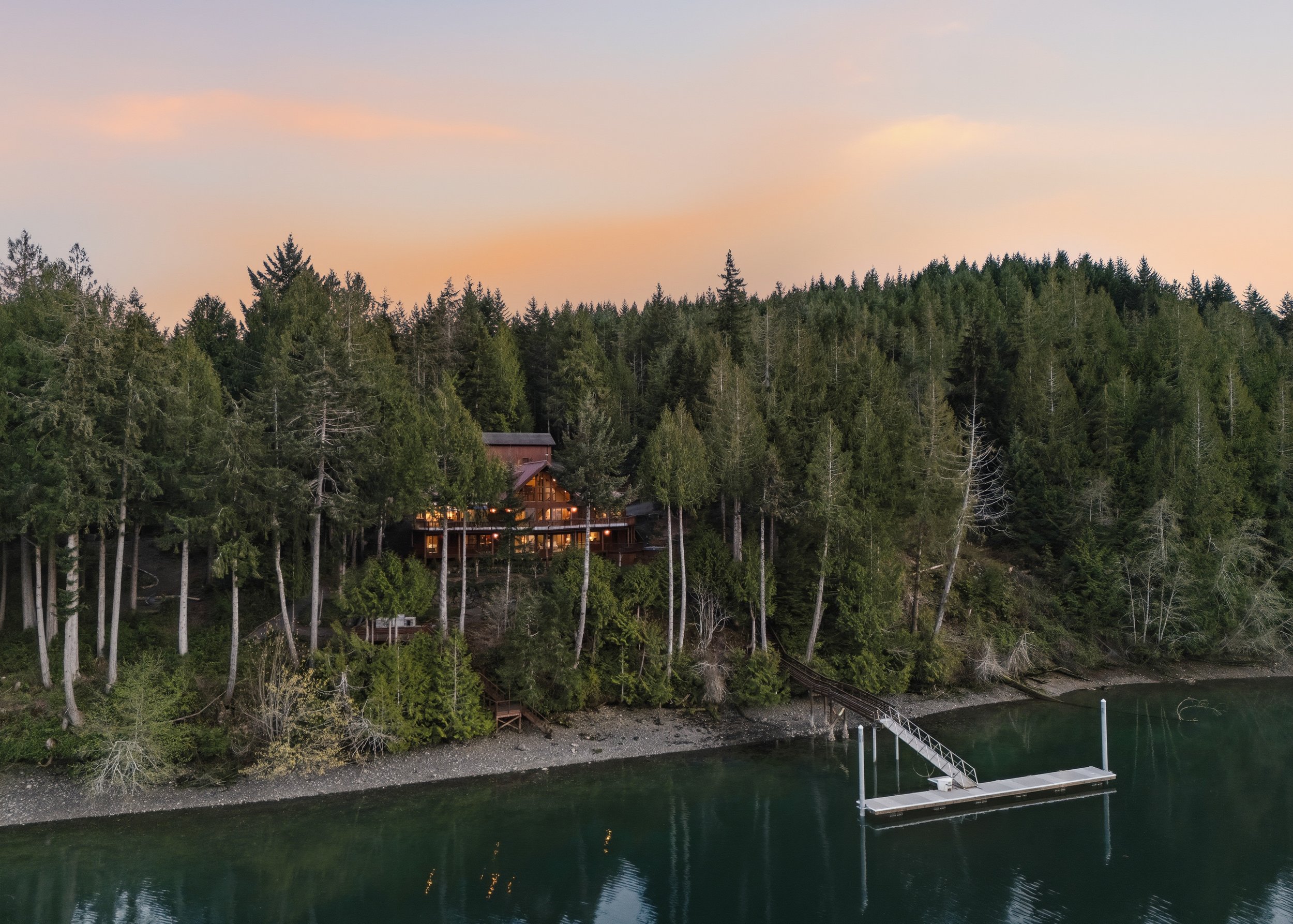 A lakeside house with lights on, surrounded by a dense forest of tall green trees, with a dock extending into calm water at sunset.