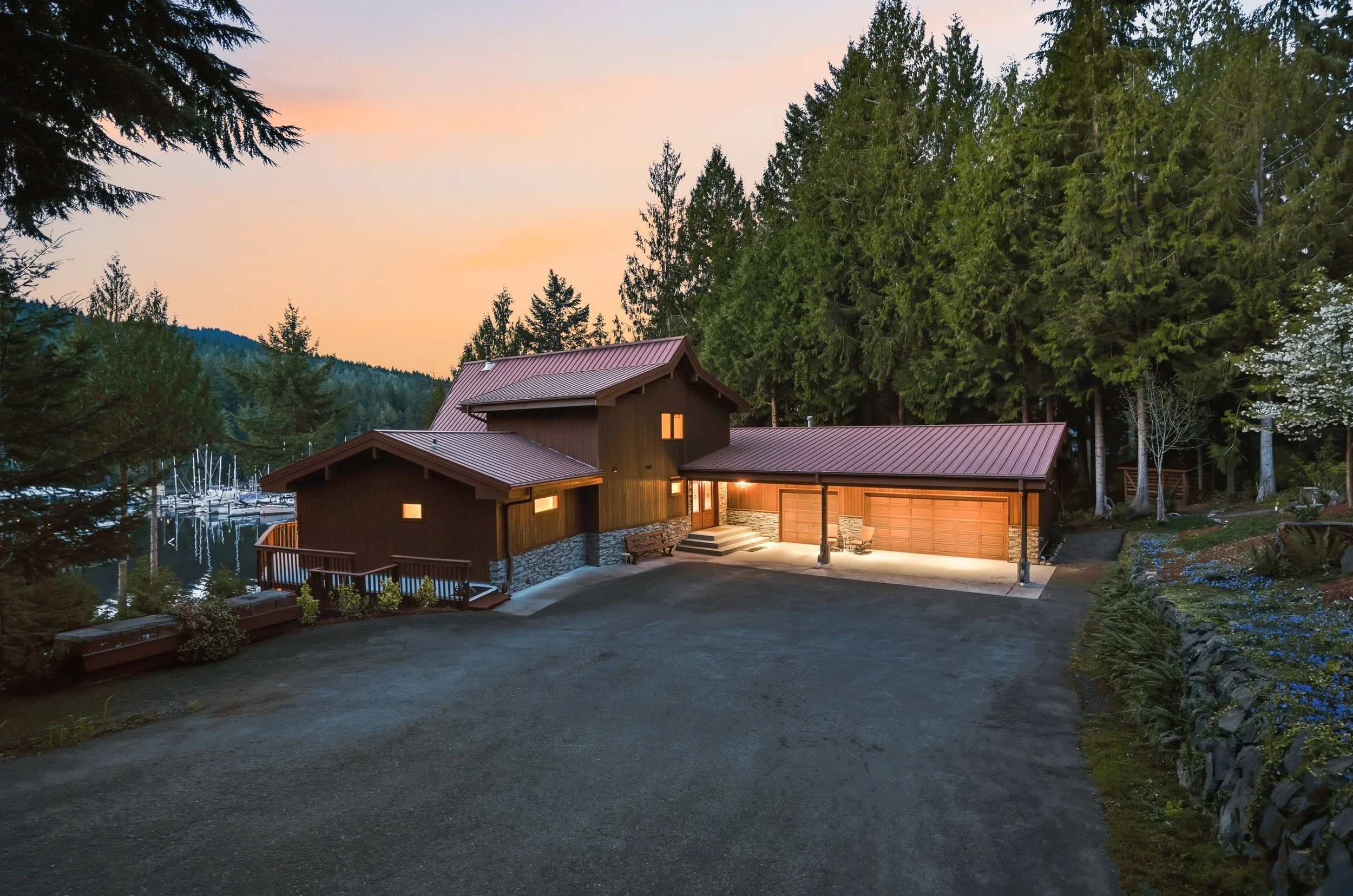 A modern house on a hillside at sunset, surrounded by trees, with a driveway leading to a garage and boat sailboats visible in the background.