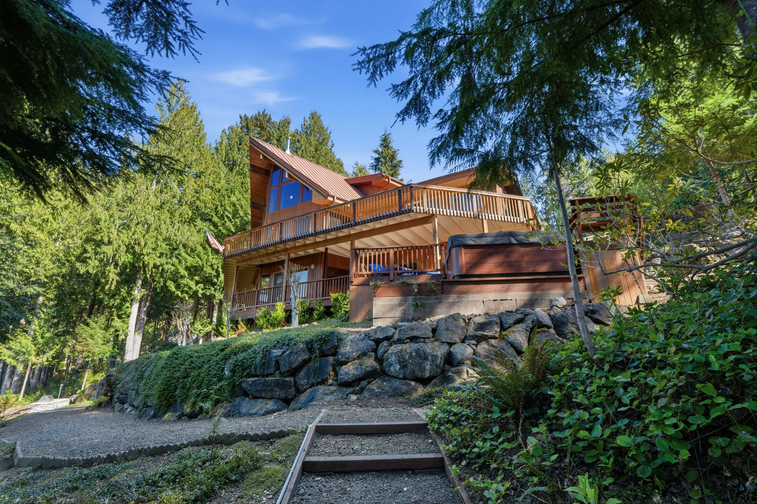 A multi-story wooden house with large windows and multiple decks, surrounded by tall green trees in a forested area.