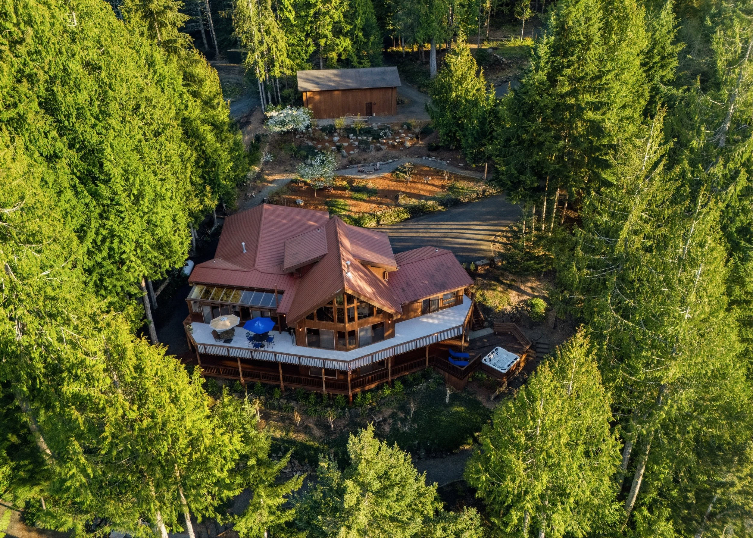 Aerial view of a large wooden house with a red roof, surrounded by tall green trees. The house features a large deck with outdoor furniture, including umbrellas, and a hot tub nearby. There is a driveway and a smaller wooden shed in the background.