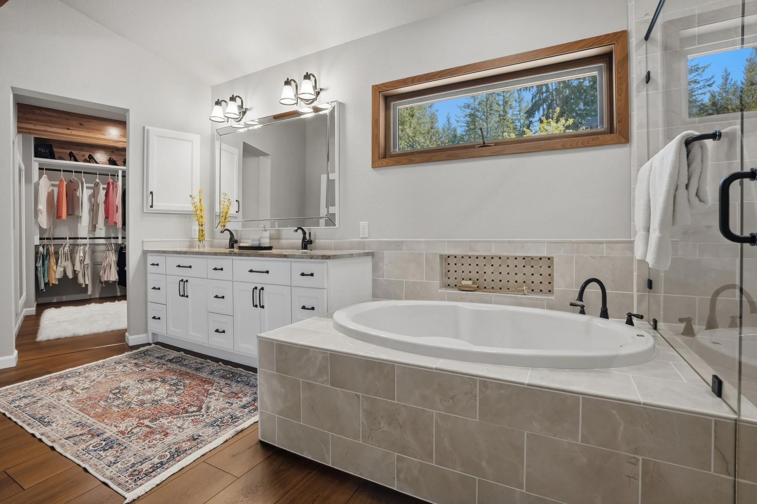 A modern bathroom with a bathtub, double vanity sink, a large mirror, and a walk-in closet with clothes and shoes visible.