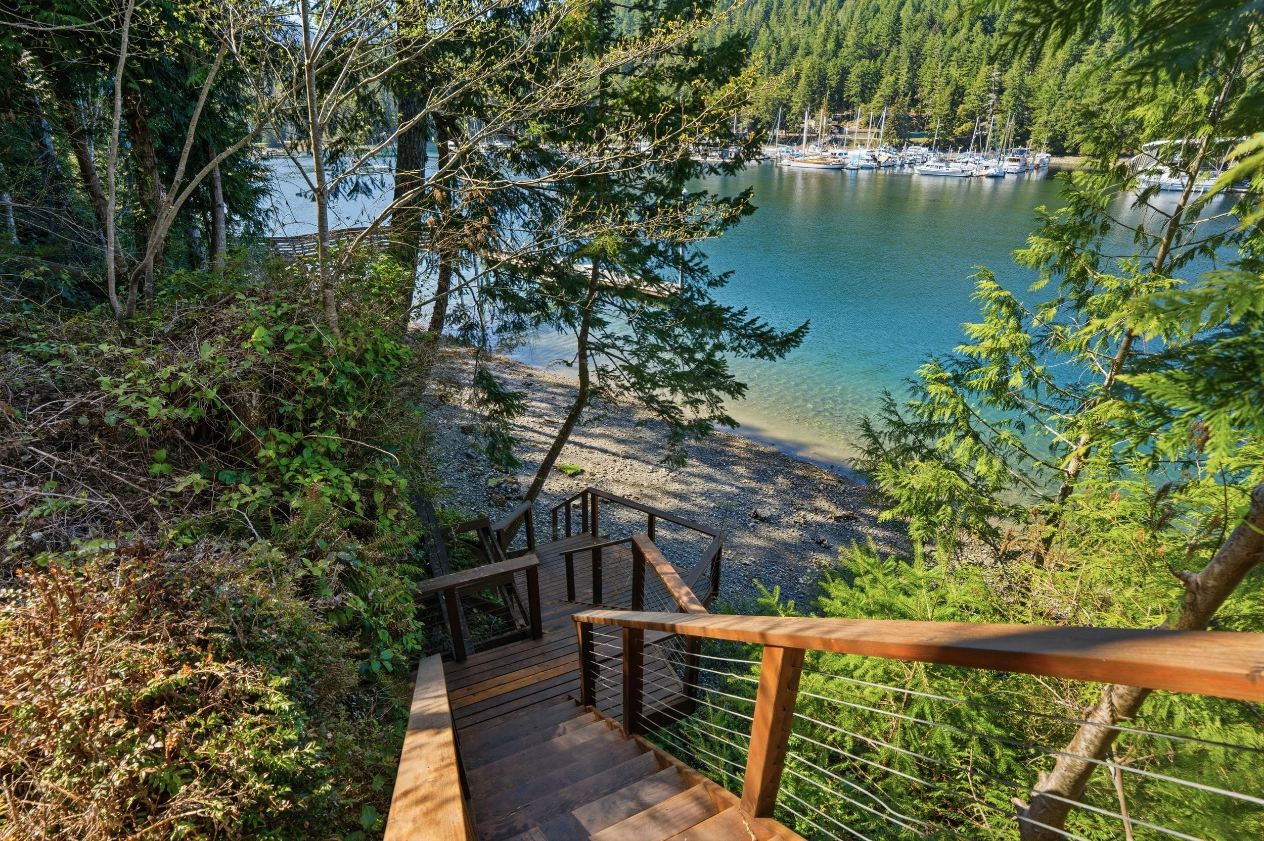 Wooden staircase leading down to a rocky beach overlooking a calm blue bay with sailboats, surrounded by dense green trees and forest.