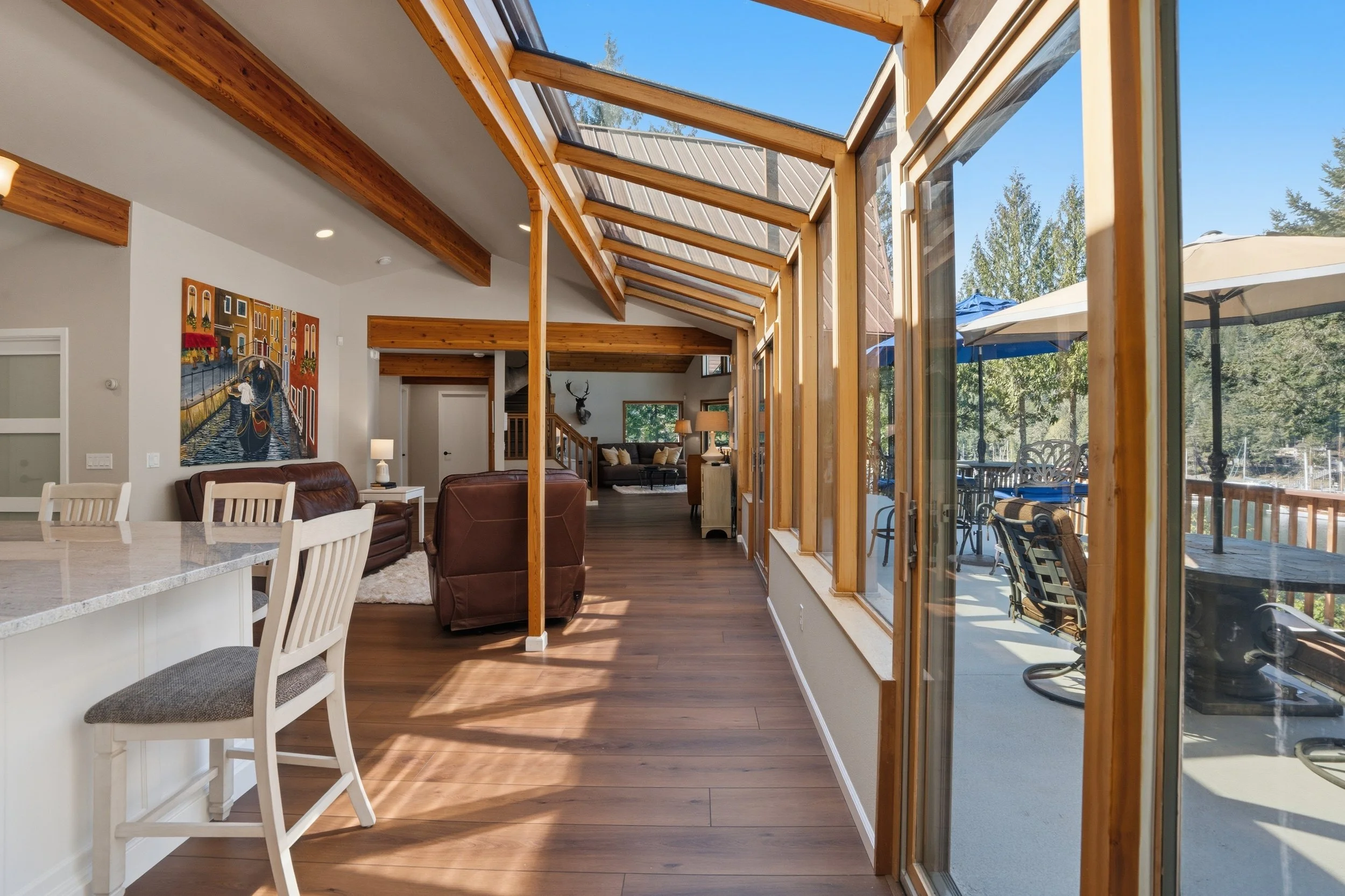 Bright living room with large windows and skylights, wooden beams, hardwood floors, and outdoor deck with patio furniture.