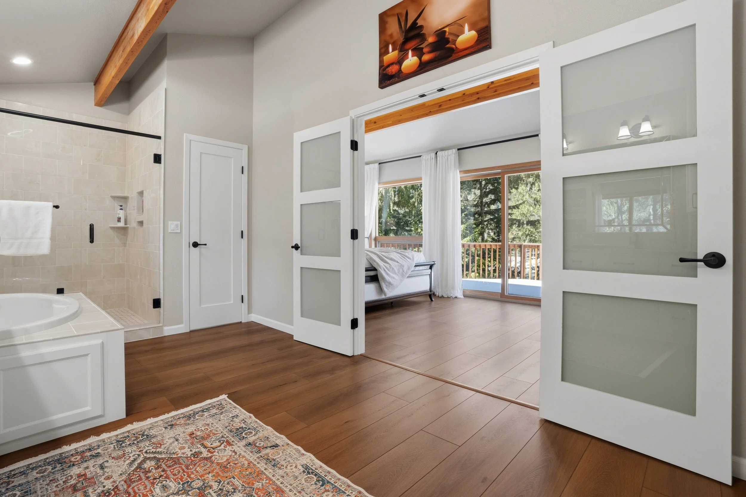 Bedroom with large windows and sliding glass door leading to balcony, white curtains, bed with white bedding, and a view of trees outside.