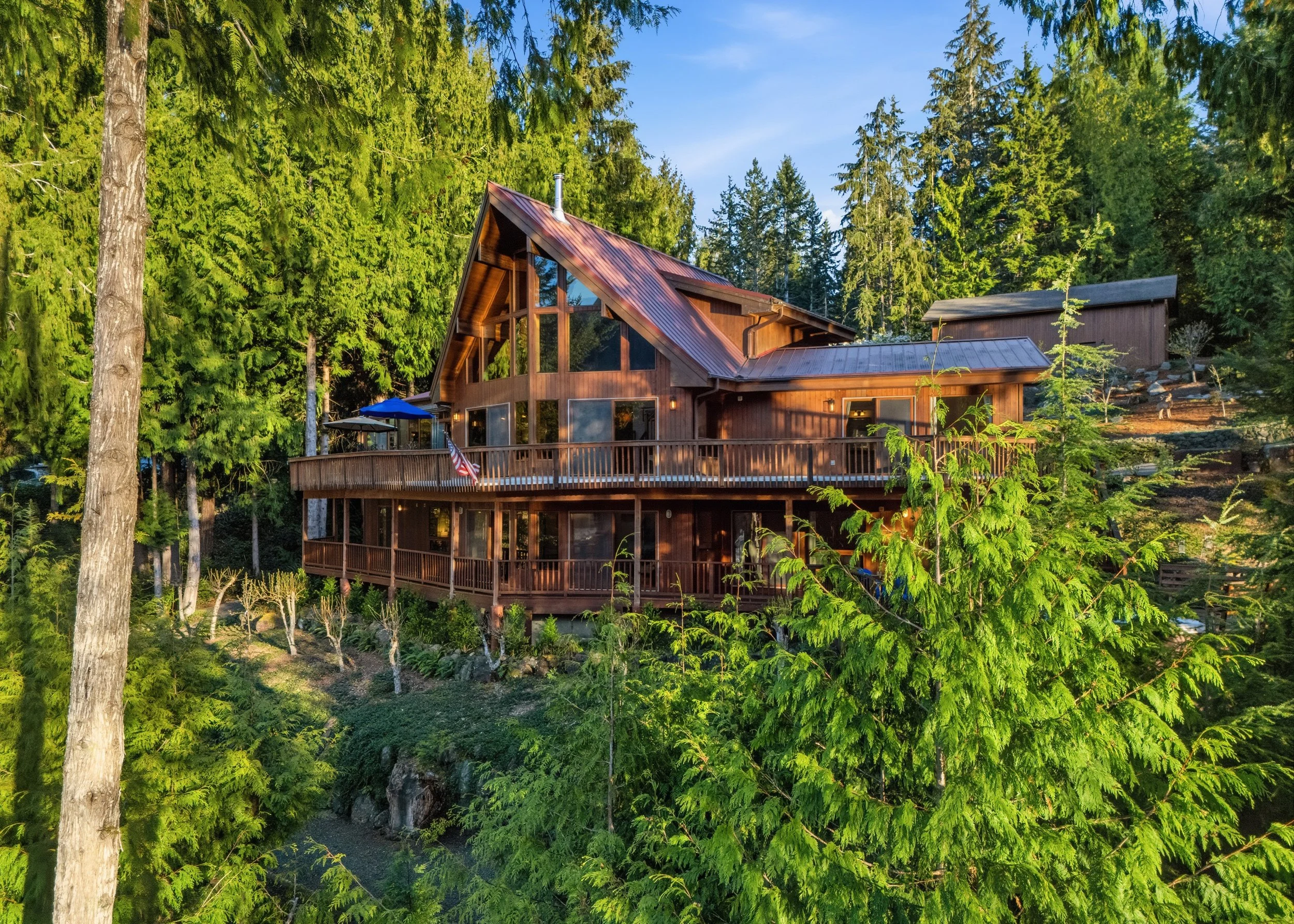 A large wooden house with a steep, triangular roof is surrounded by tall green trees and foliage. The house features expansive decks, large windows, and an American flag hanging from one side.