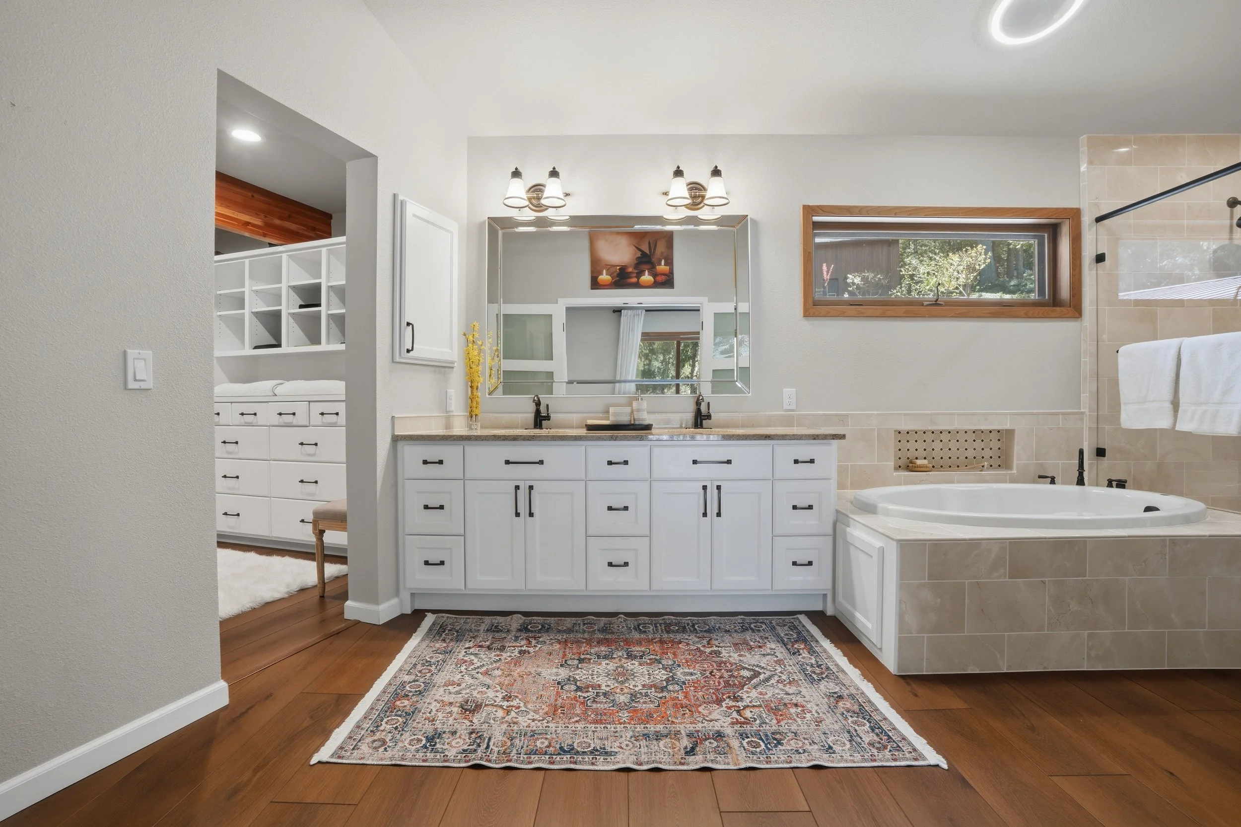 A modern bathroom with a white vanity, double sinks, large mirror, beige tile bathtub surround, window, and a decorative rug on wooden floor.