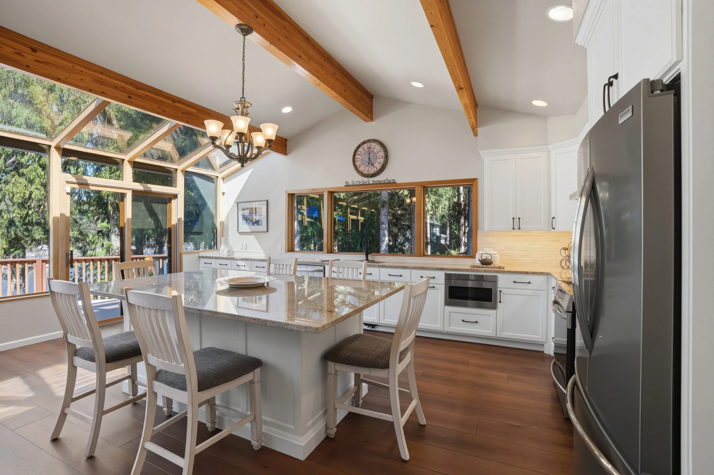 Bright, spacious kitchen with large window bay and exposed wooden ceiling beams, white cabinetry, gray countertop, stainless steel appliances, and a central island with seating.