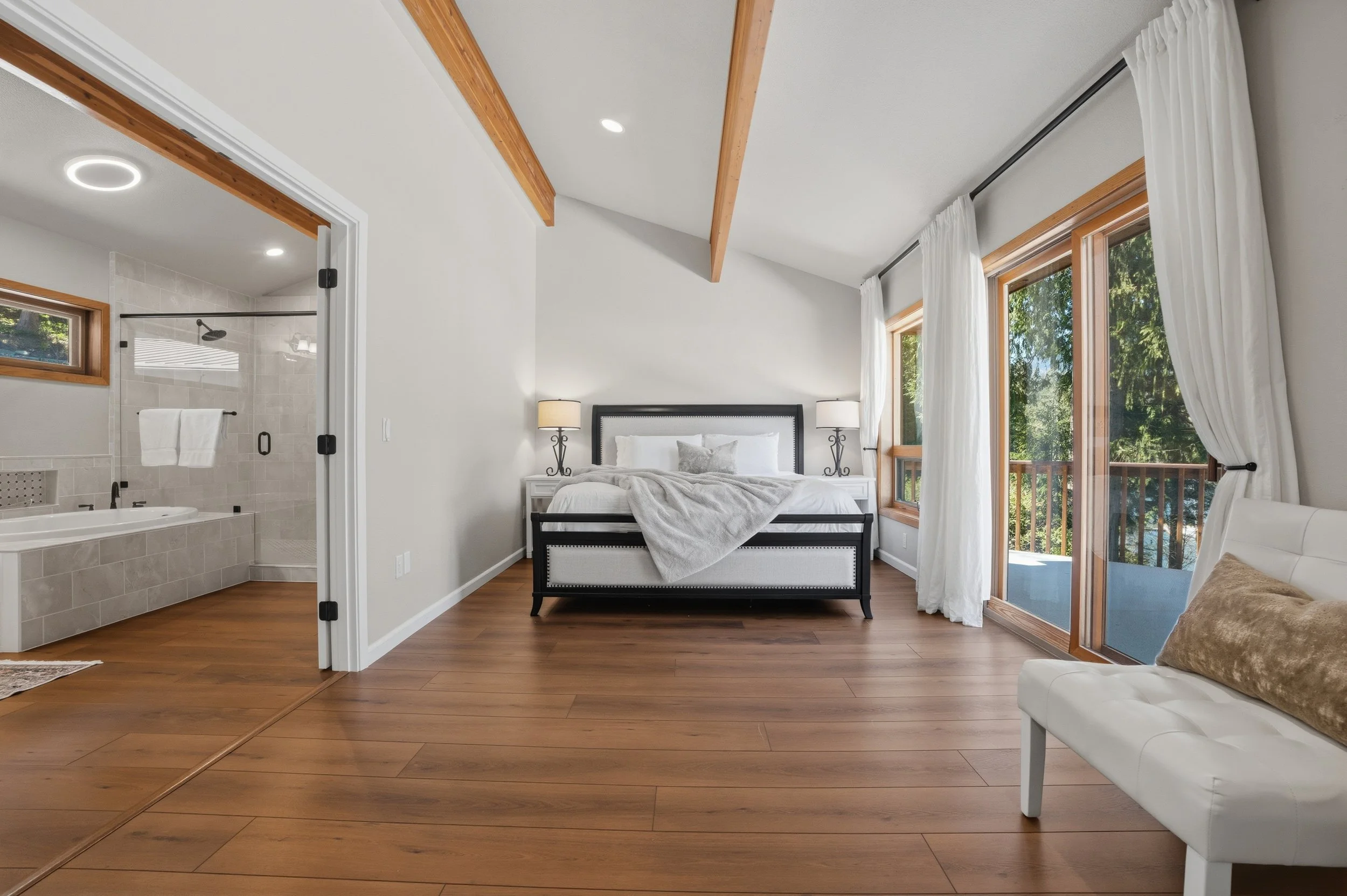Bedroom with large windows, white curtains, and a view of trees outside. A bed with white bedding and gray pillows is flanked by two lamps. A white bench with a gold pillow is visible on the right. The bathroom with a bathtub and shower is visible th