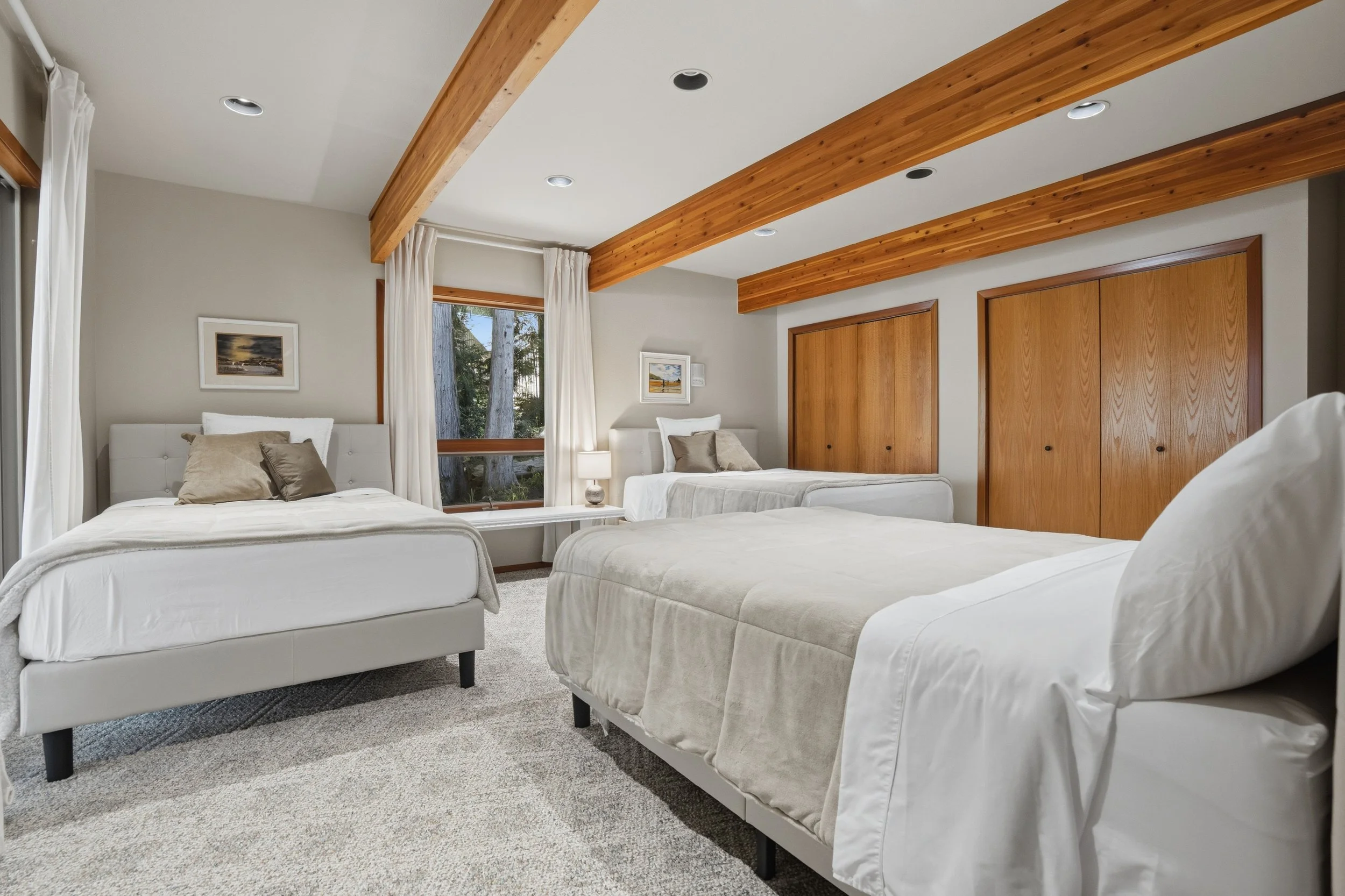 A bedroom with three beds, white bedding, beige and gray pillows, wooden beams on the ceiling, window with white curtains, and two framed pictures on the wall.
