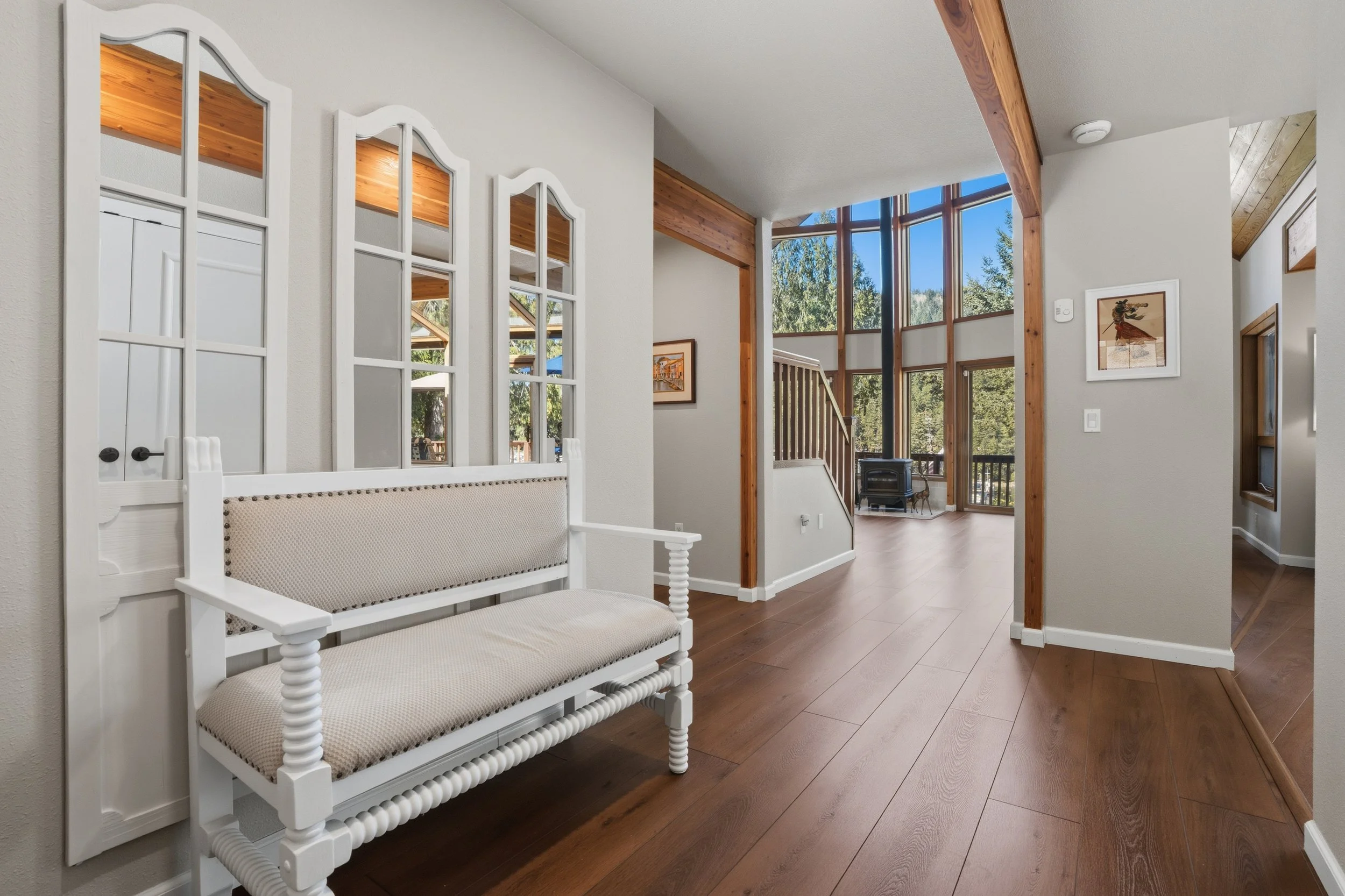 Entryway with wooden floors, a white bench, decorative windows, and a view into a sunlit room with large windows and a staircase.