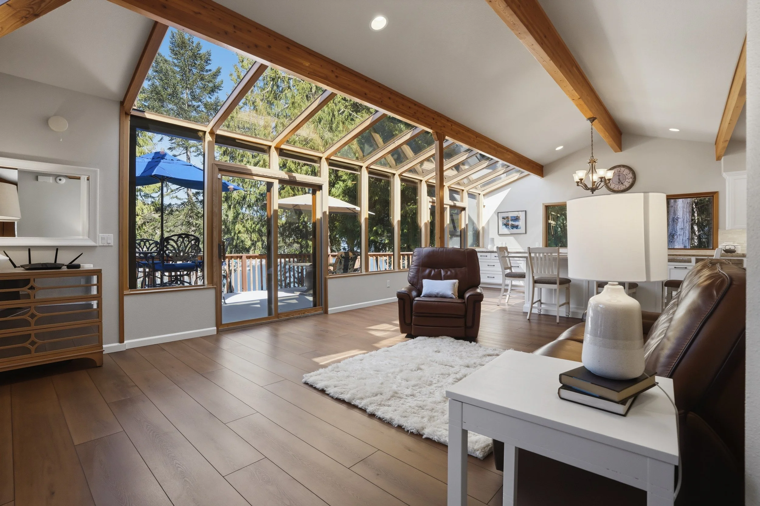 Living room with large glass window and door leading to outdoor deck, featuring wooden beams on ceiling, leather armchair, white side table with lamp, and hardwood floors.