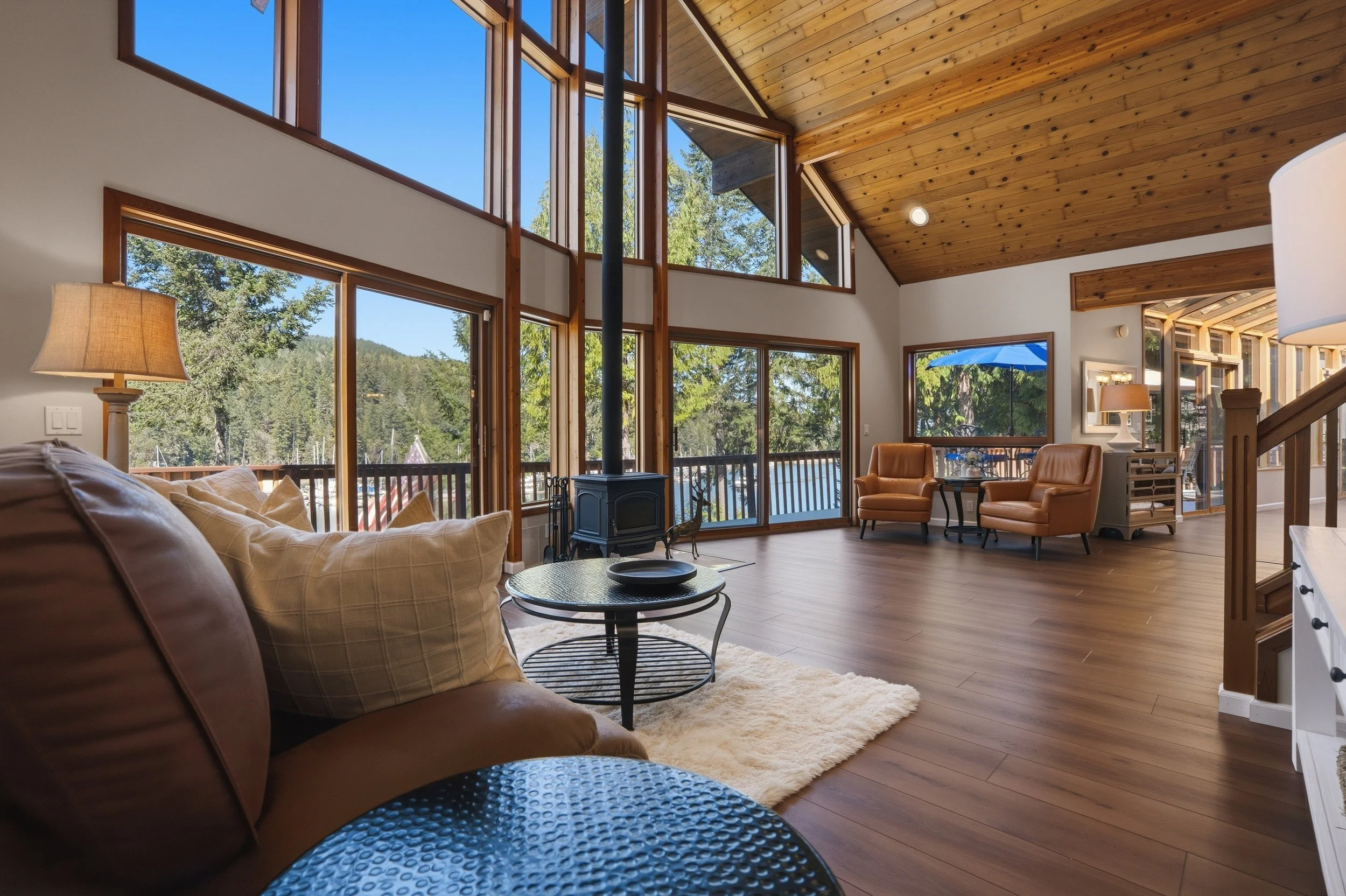 Spacious living room with large wooden-framed windows and skylights, exposed wooden ceiling, hardwood floors, sofa with pillows, leather chairs, side table, wood stove, and backyard view with trees and mountains.