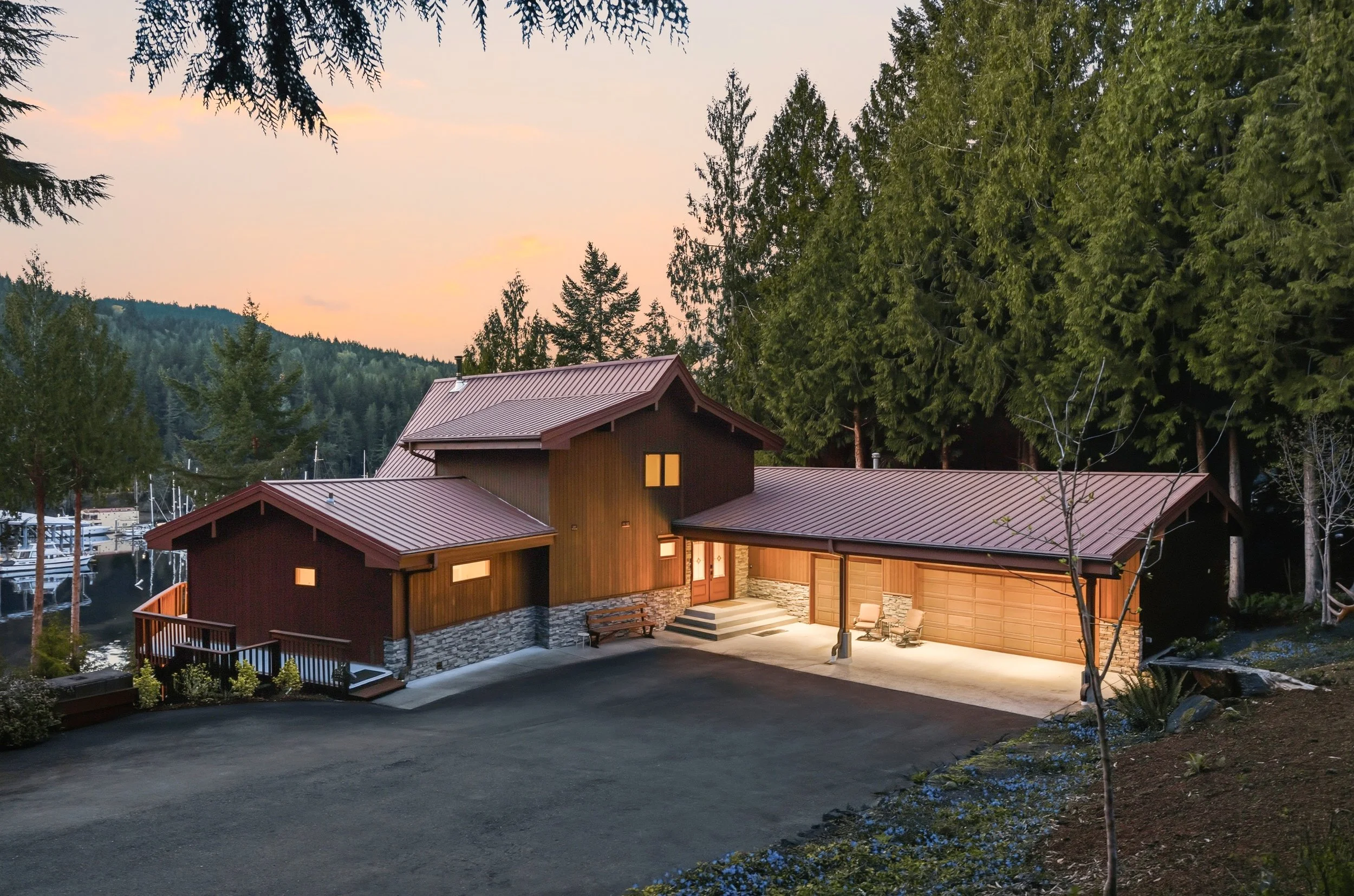 A modern house with a red metal roof, stone accents, and large garage doors in a wooded area during dusk.