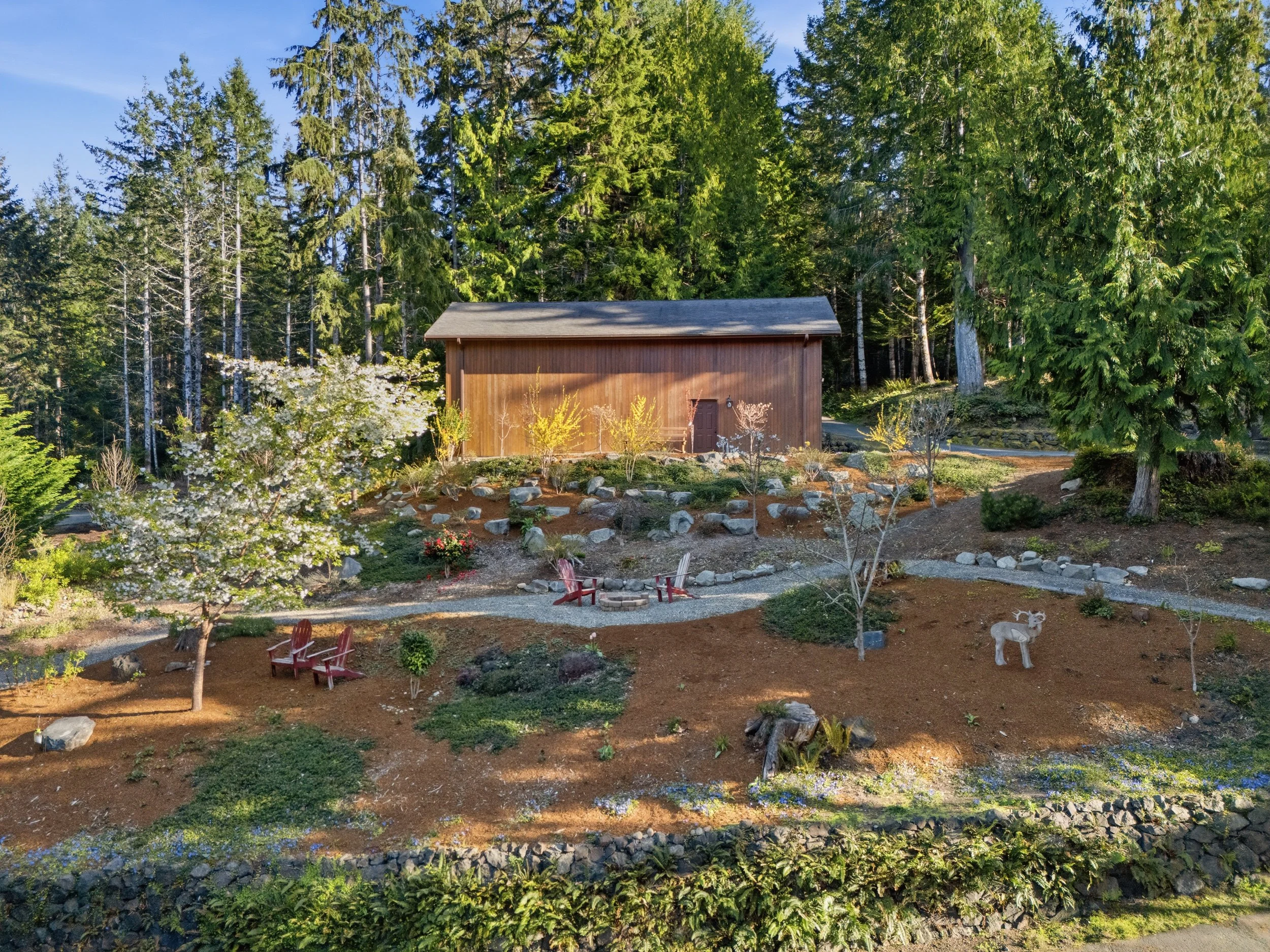 A landscaped backyard with trees, a wooden shed, and garden furniture, including red Adirondack chairs, set in a forested area with large trees and a gravel pathway.