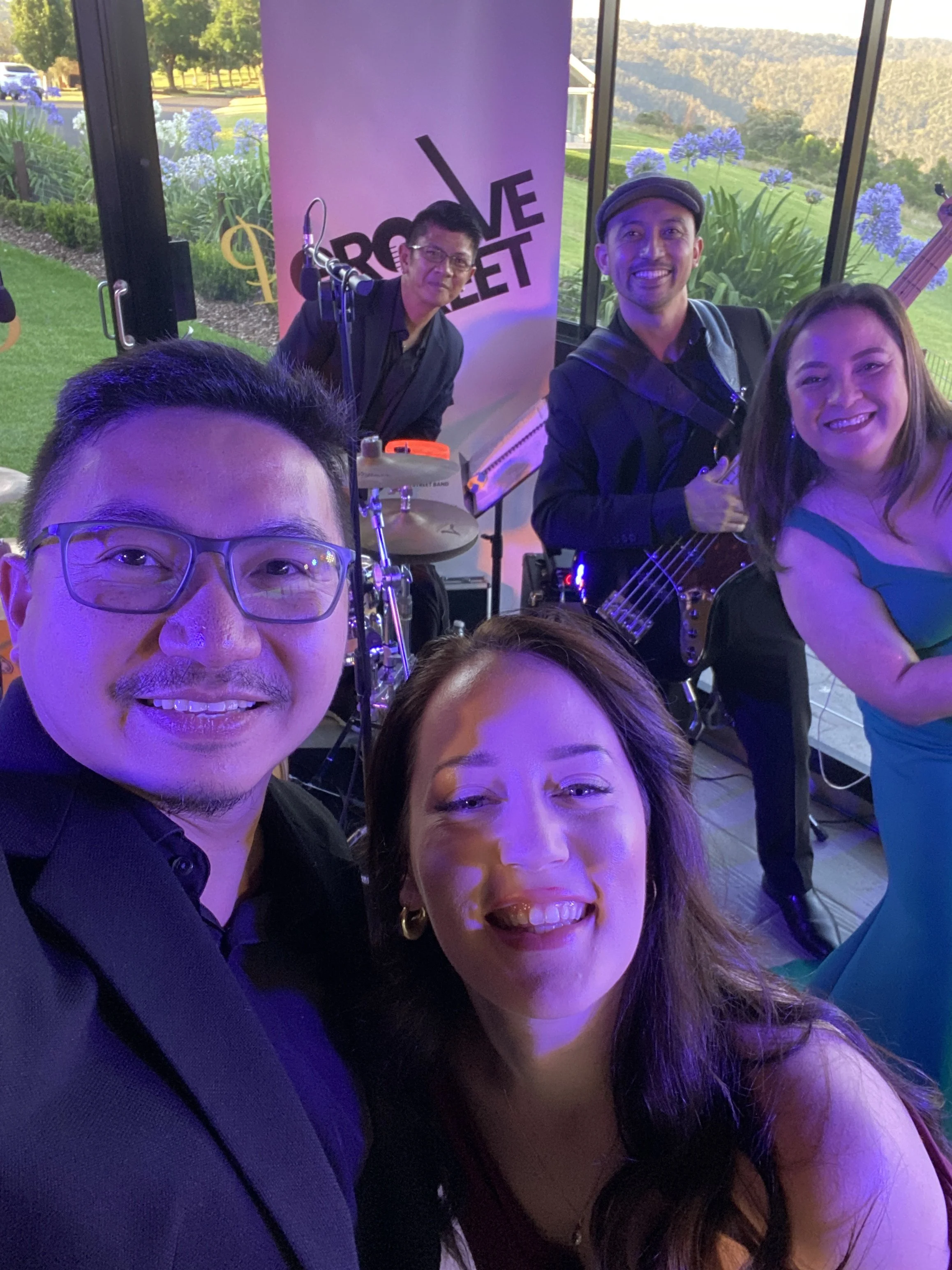 A group of five people, smiling and posing for a selfie during a live music performance indoors with large windows showing a garden and hills outside. The band members are playing instruments, including drums, bass guitar, and keyboard. A banner behi
