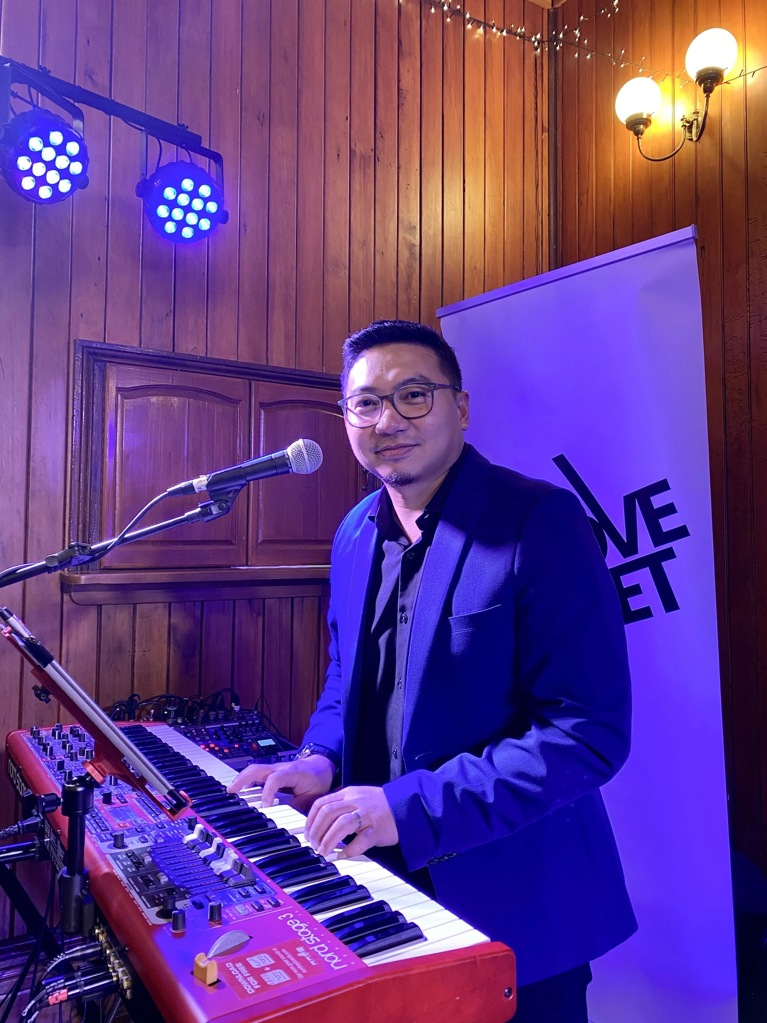 A man wearing glasses and a dark blue blazer playing a red keyboard in a wooden room with purple lighting, a microphone in front of him, and a white banner with black text in the background.