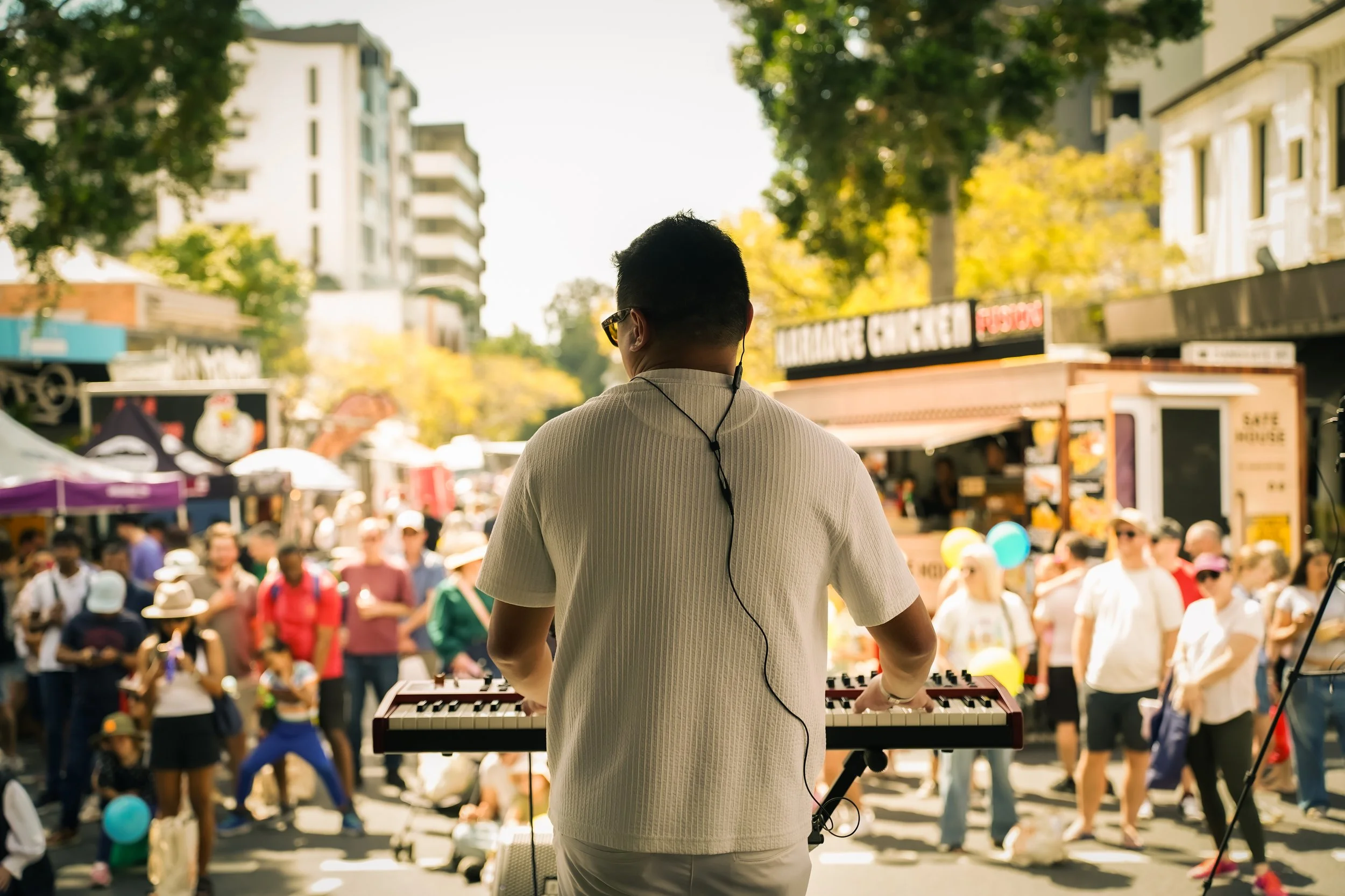 A man playing a keyboard at an outdoor market or event with a crowded crowd in the background.
