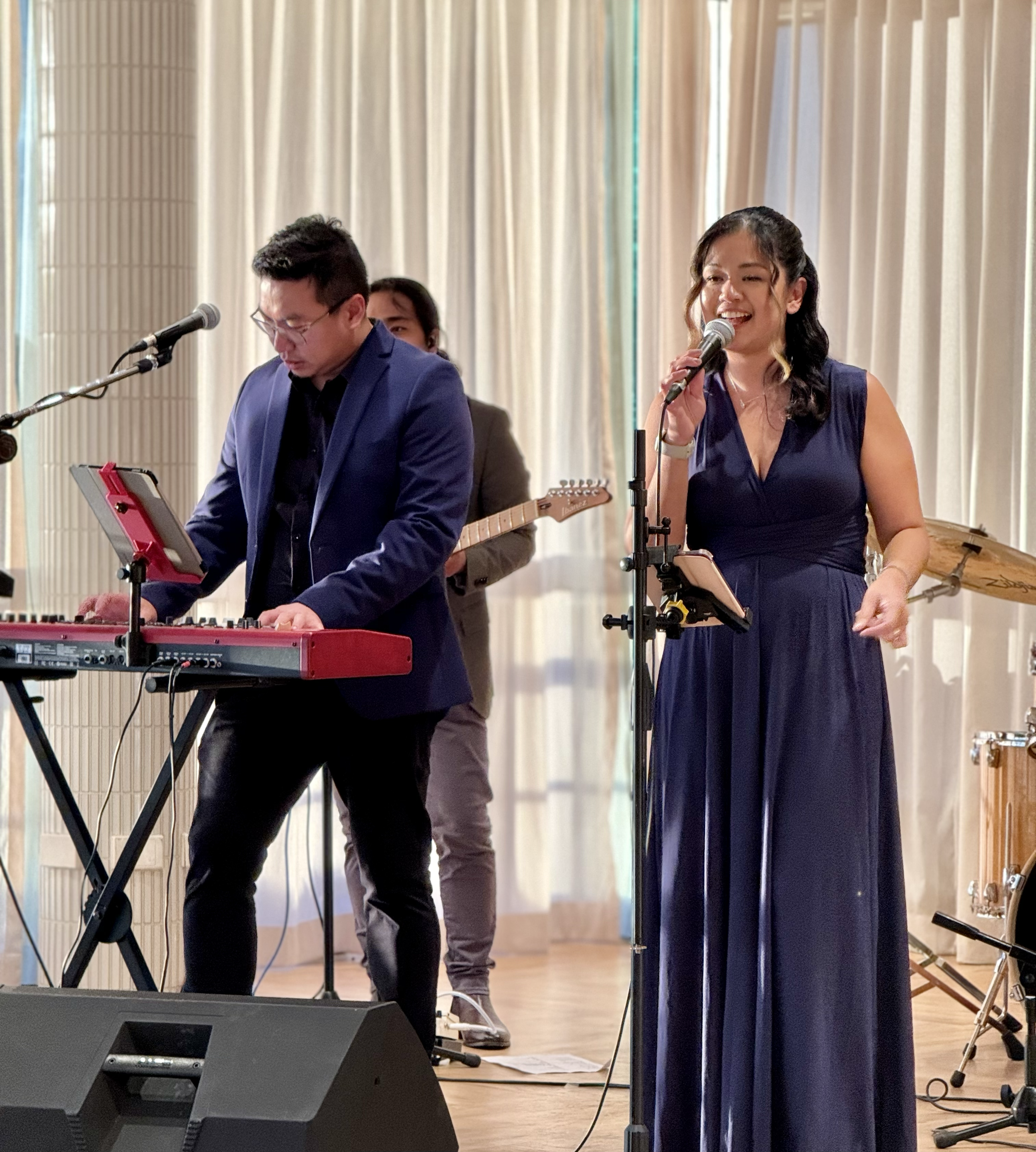Musicians performing on stage: a woman in a navy blue dress singing into a microphone, a man in a navy blazer playing a keyboard, and a person in the background playing guitar, with a drum set nearby.