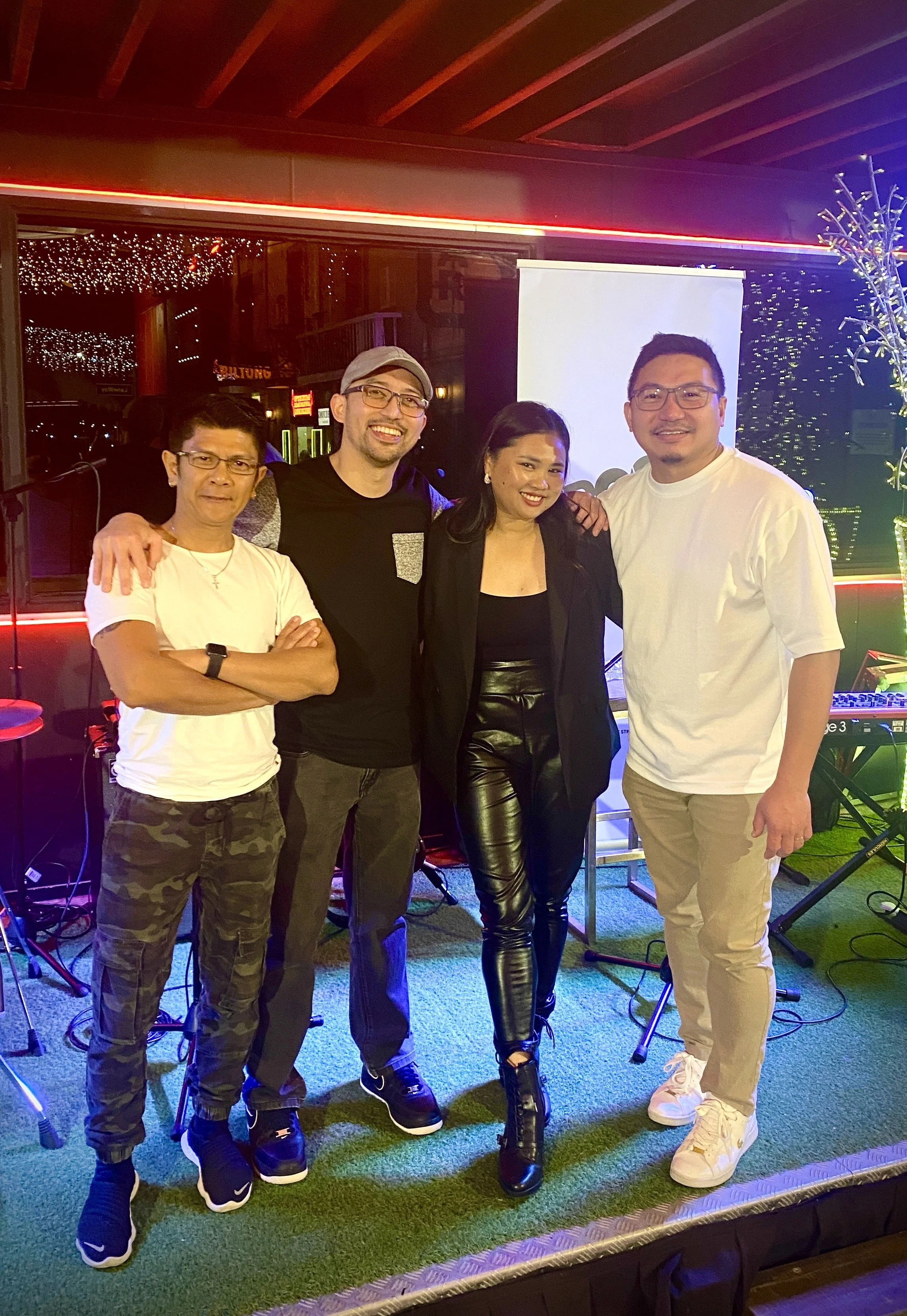Group of four people standing together at an indoor event with musical equipment and a projection screen in the background, smiling for the camera.