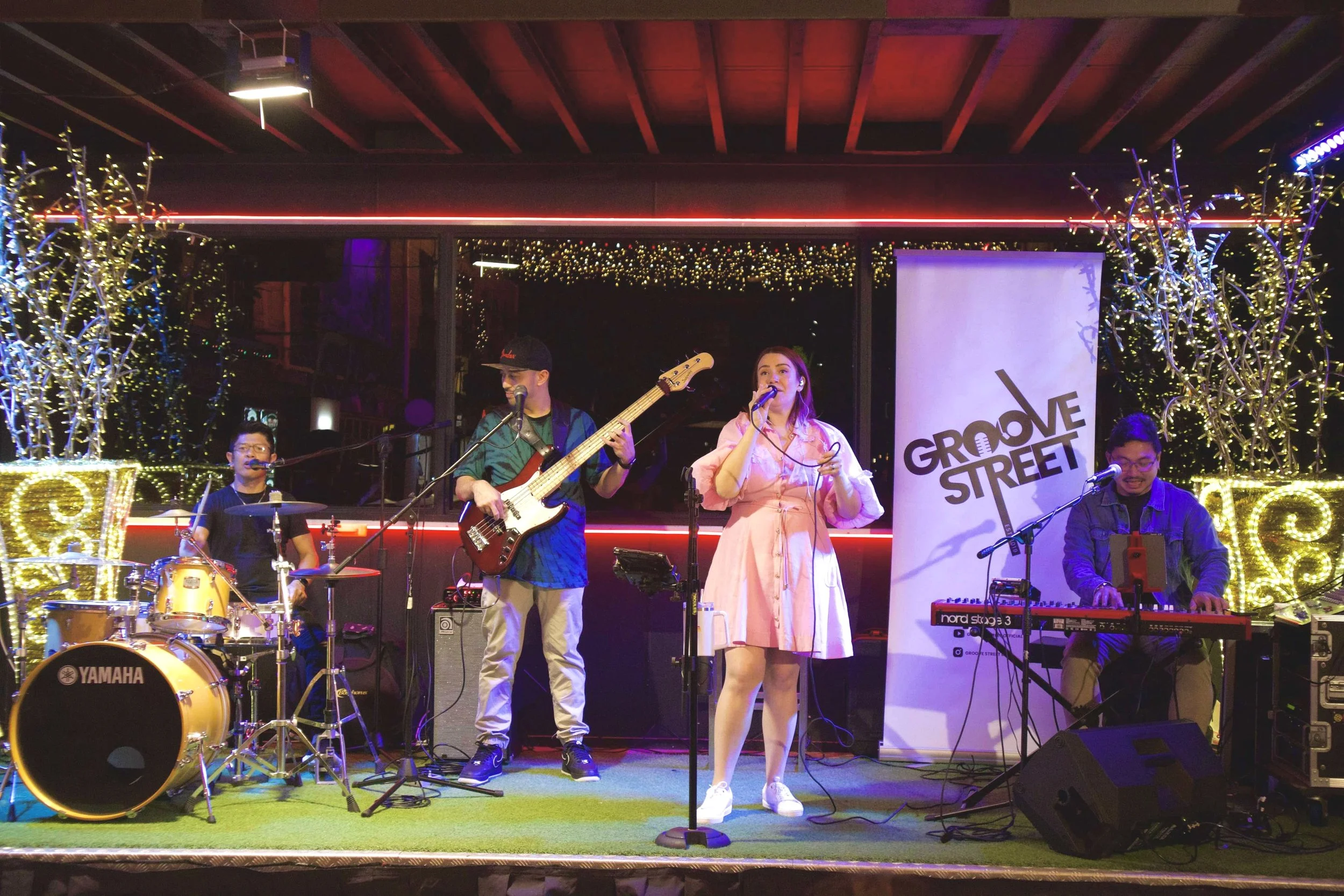 A band performing live on stage with four musicians, including a drummer, bassist, singer, and keyboard player, illuminated by colorful stage lights, with a 