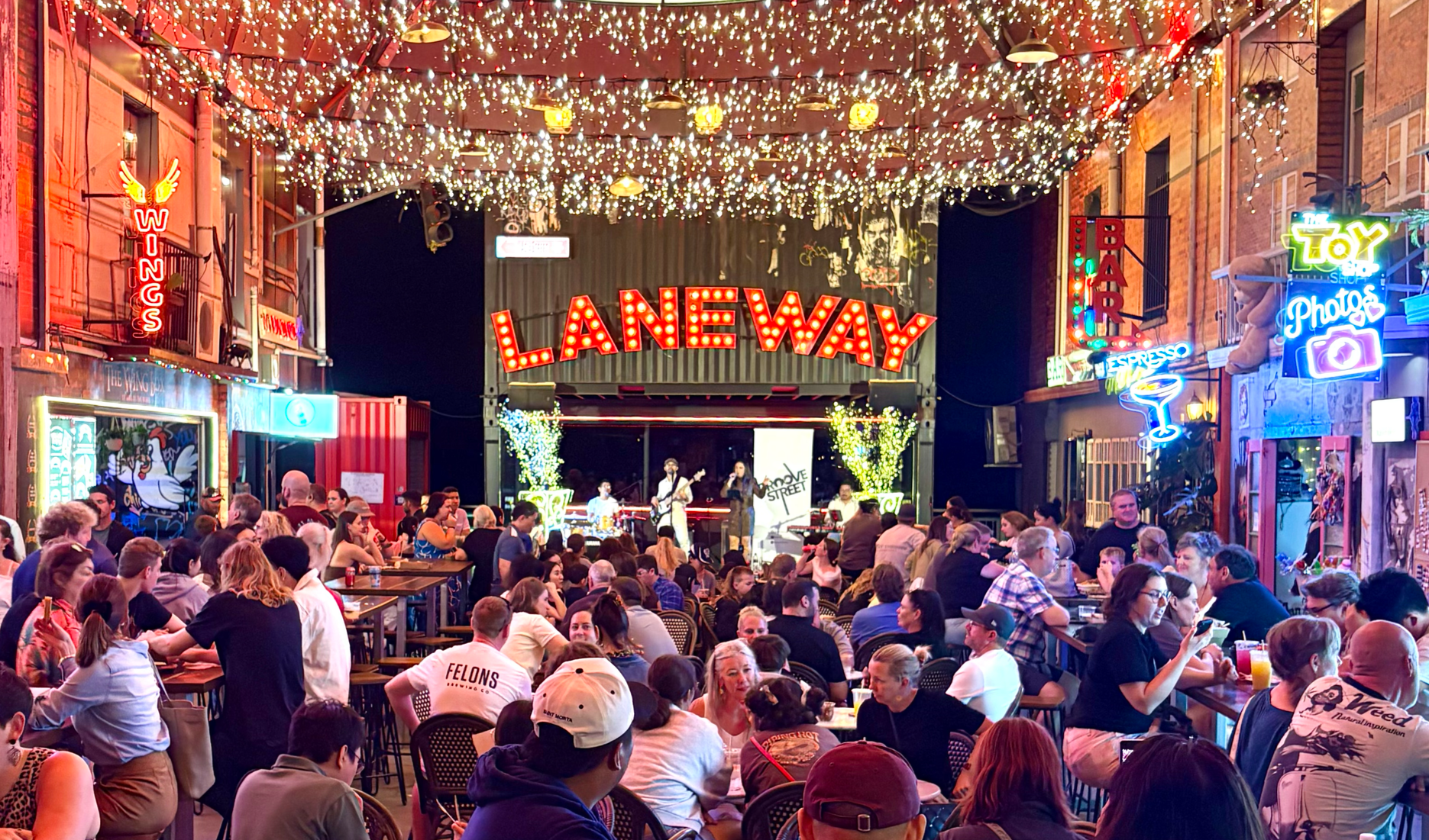Indoor venue with a stage hosting live music, surrounded by a crowd sitting at tables. The ceiling is decorated with many string lights. Bright neon signs on the walls include 'WINGS', 'BAR', and 'TOY Photos'. A large red marquee sign says 'LANEWAY'.