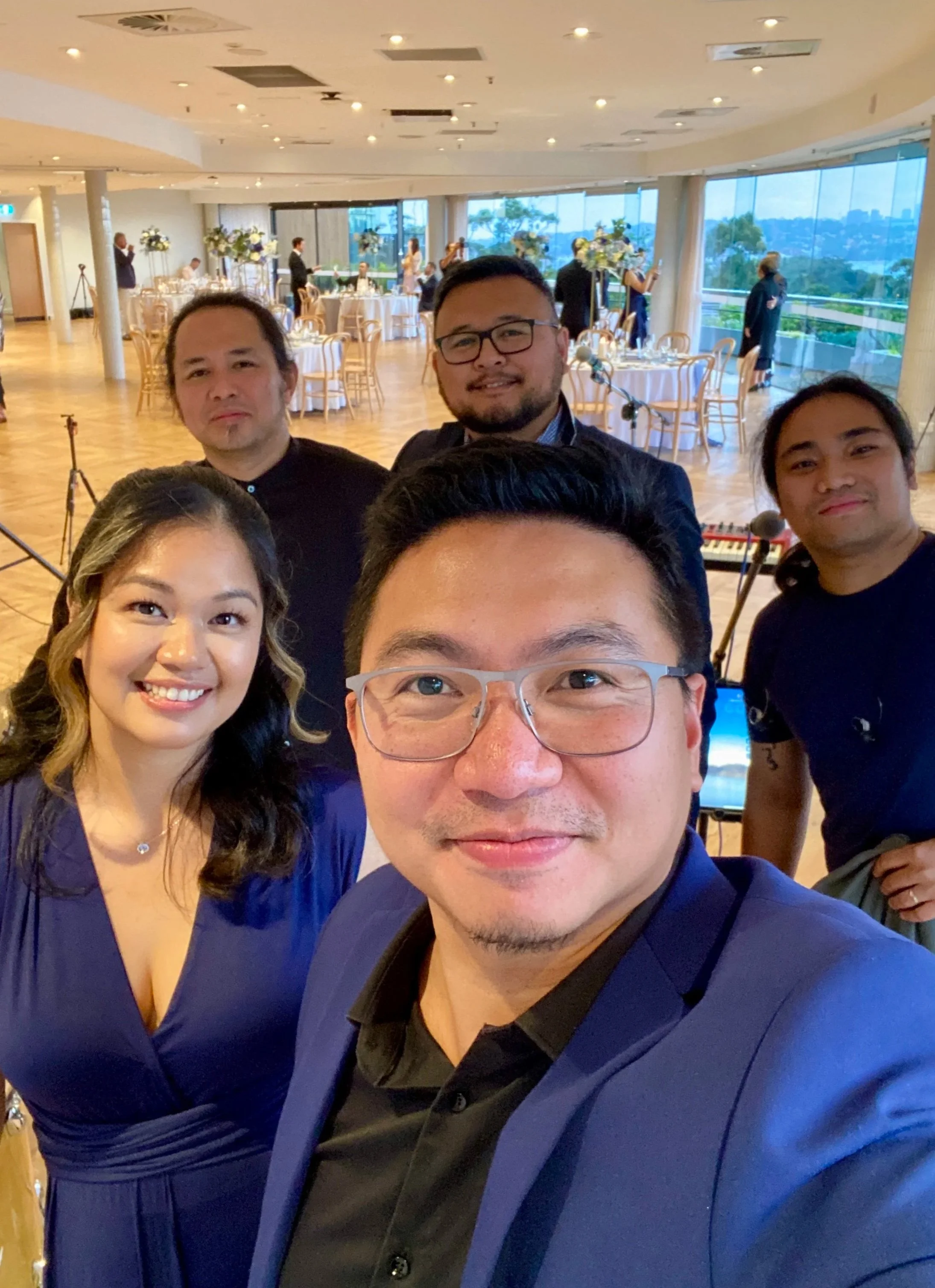 Group of six people taking a selfie at a formal event in a decorated venue with large windows and round tables.