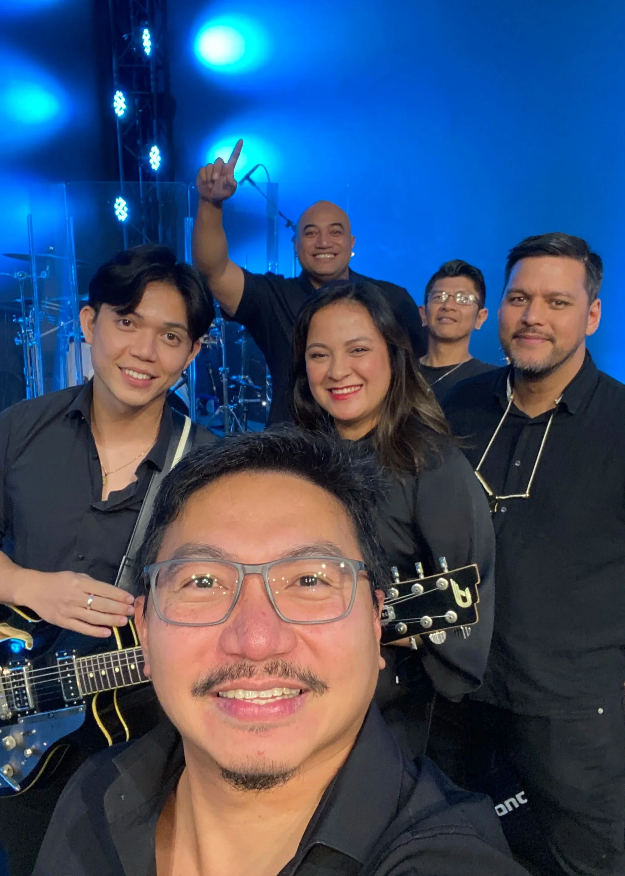 Group of six people on stage or a concert venue, smiling and posing for a selfie. The person in the foreground is wearing glasses and holding a guitar, while the others are standing behind him, some with glasses and some smiling and looking at the ca