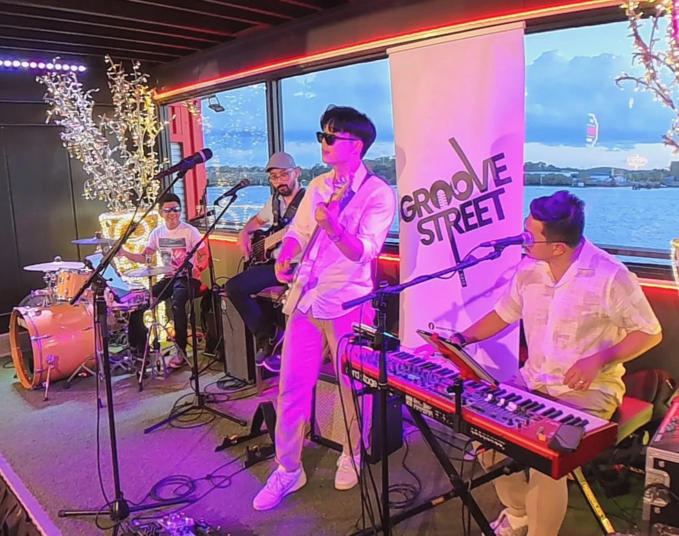 A live band performing in a room with large windows overlooking a river. The band includes a drummer, a guitar player, a bassist, and a keyboardist. The scene is colorful with pink and purple lighting, and the band members are wearing casual clothing