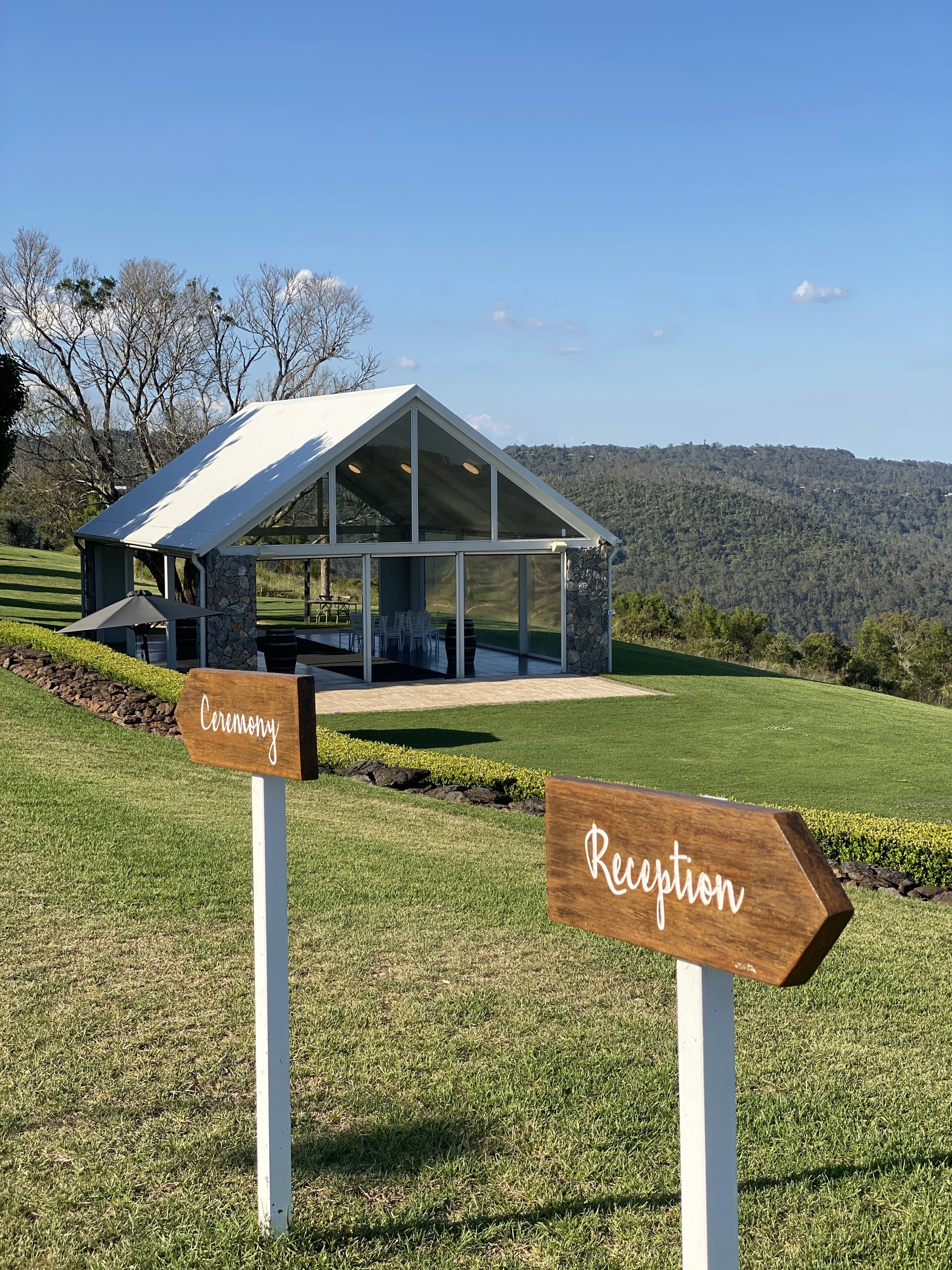 An outdoor wedding venue with a modern glass and stone pavilion on a grassy hill, surrounded by trees and mountains in the background. There are signs in the foreground pointing to the 'Ceremony' and 'Reception' areas.