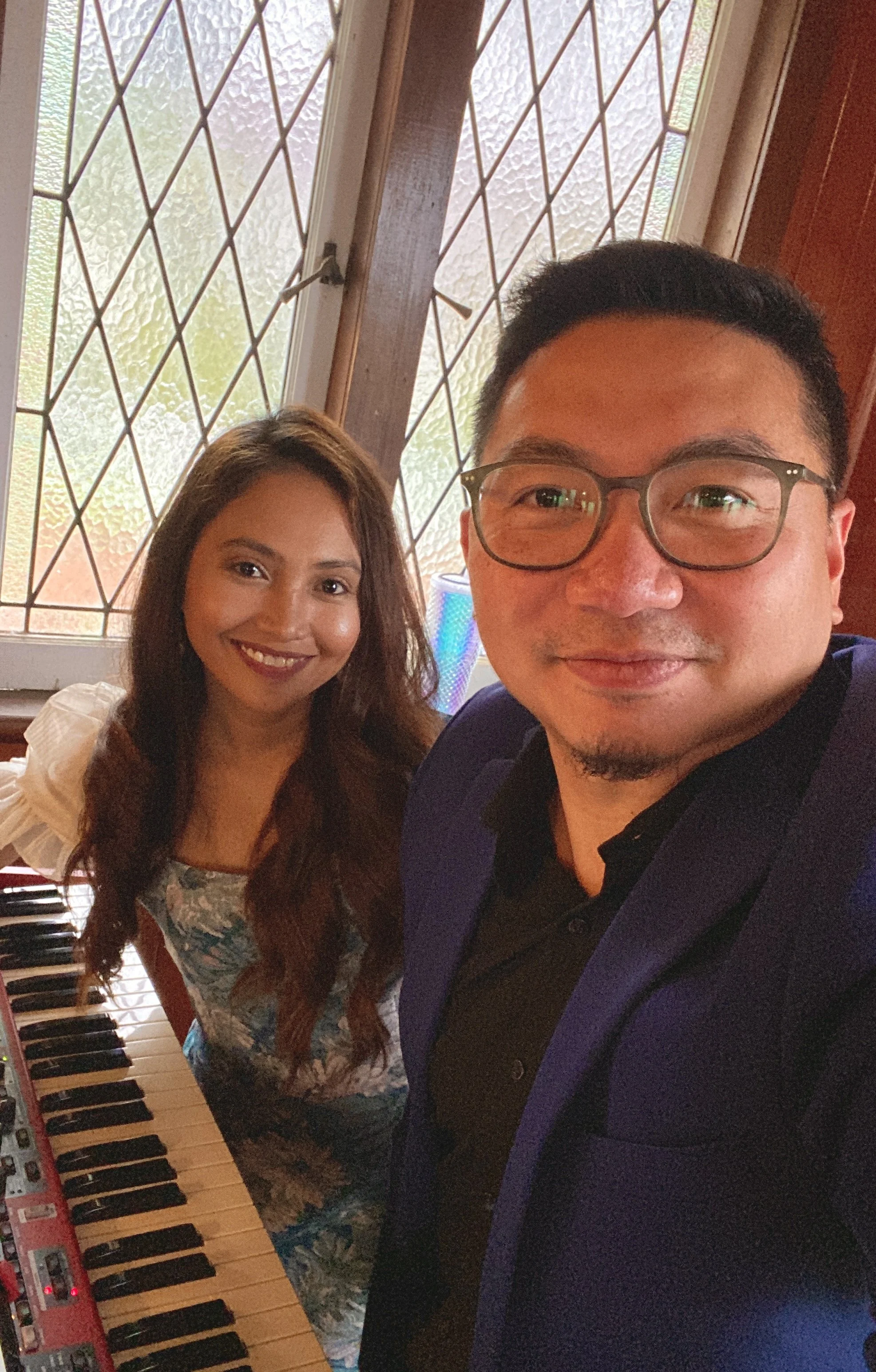 A man and a woman smiling for a selfie indoors near a window and a keyboard instrument.