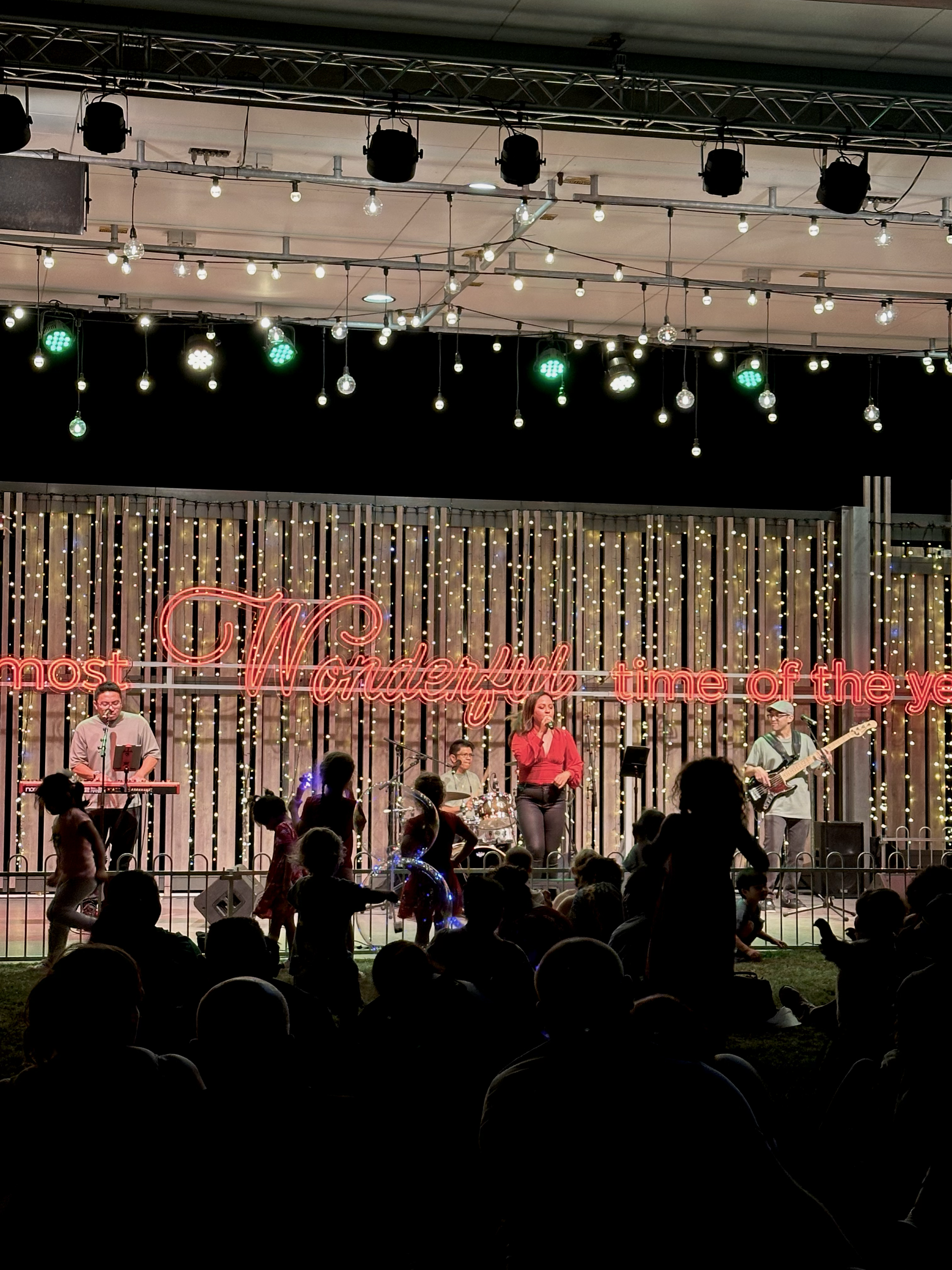 A live music performance on an outdoor stage at night, with a band including a keyboardist, drummer, female singer, and bass guitarist. The stage is decorated with string lights and a neon sign reading 'Wonderful.' Audience members are sitting and st