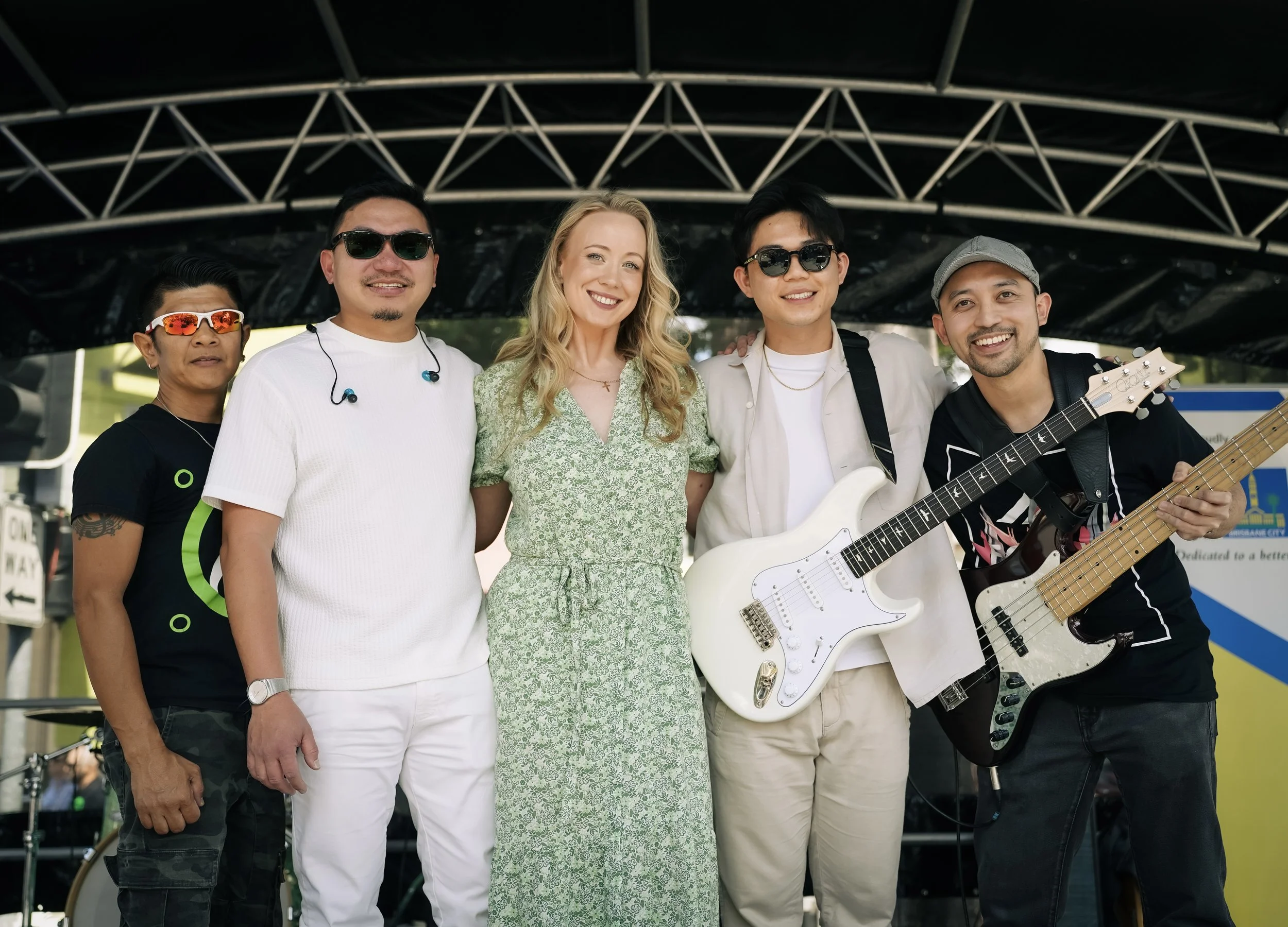 Group of five diverse musicians and a woman smiling, standing on a stage with musical instruments, outdoor setting.